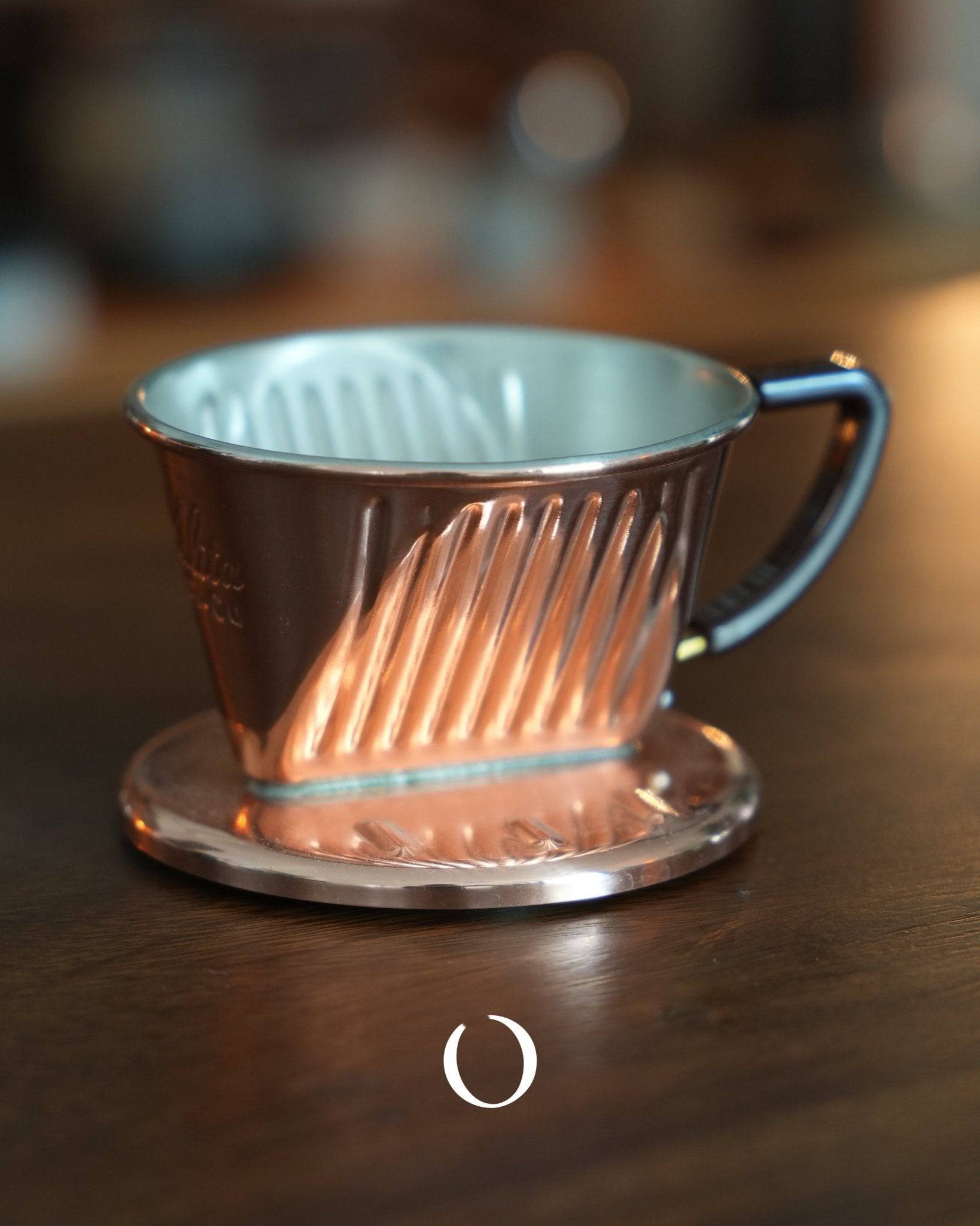KALITA copper pour-over coffee dripper with three-hole design, featuring a conical shape and ribbed interior, sitting on a metal base, showcasing its handcrafted quality and unique copper finish for optimal heat distribution and balanced extraction