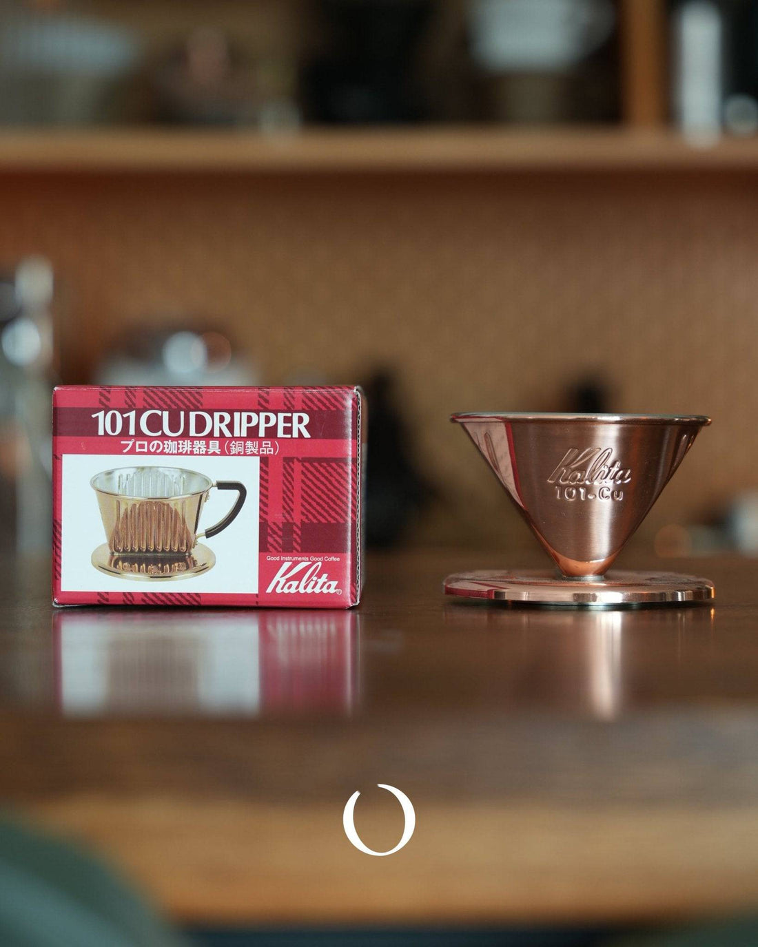 Kalita 101-CU copper coffee dripper with three-hole design, displayed next to its red packaging box on a wooden surface. The dripper&