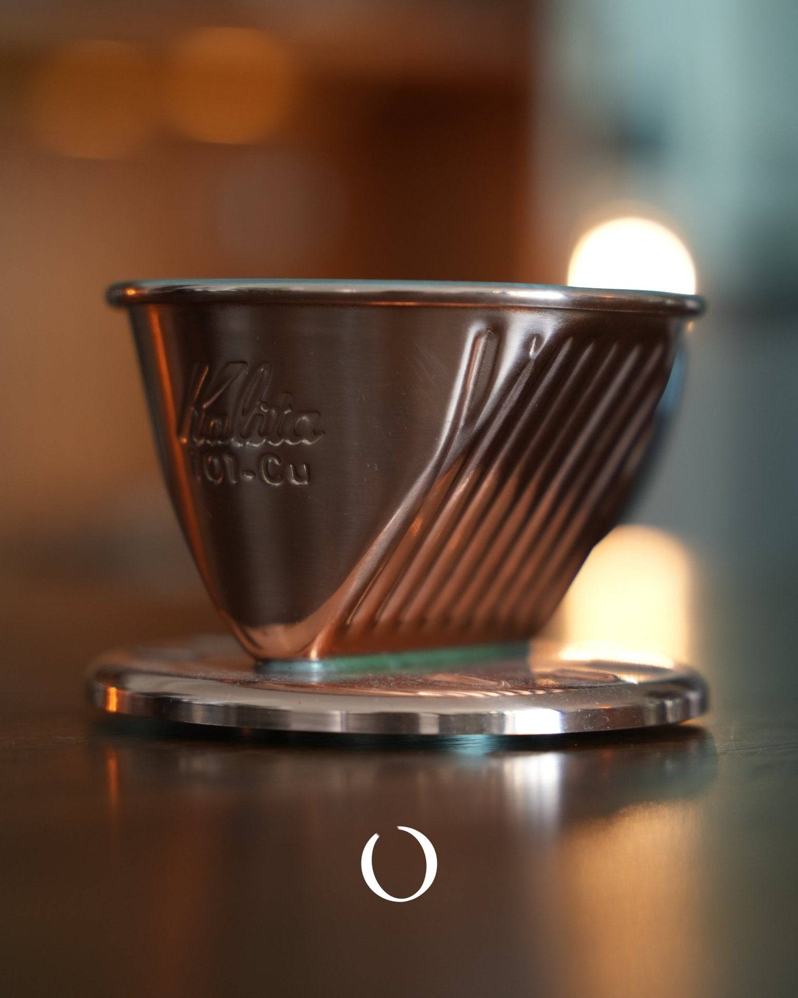 Kalita 101-CU copper coffee dripper with three-hole design, showcasing its unique conical shape and ribbed exterior, positioned on a reflective surface with warm background lighting, highlighting its premium craftsmanship and aesthetic appeal for coffee enthusiasts.