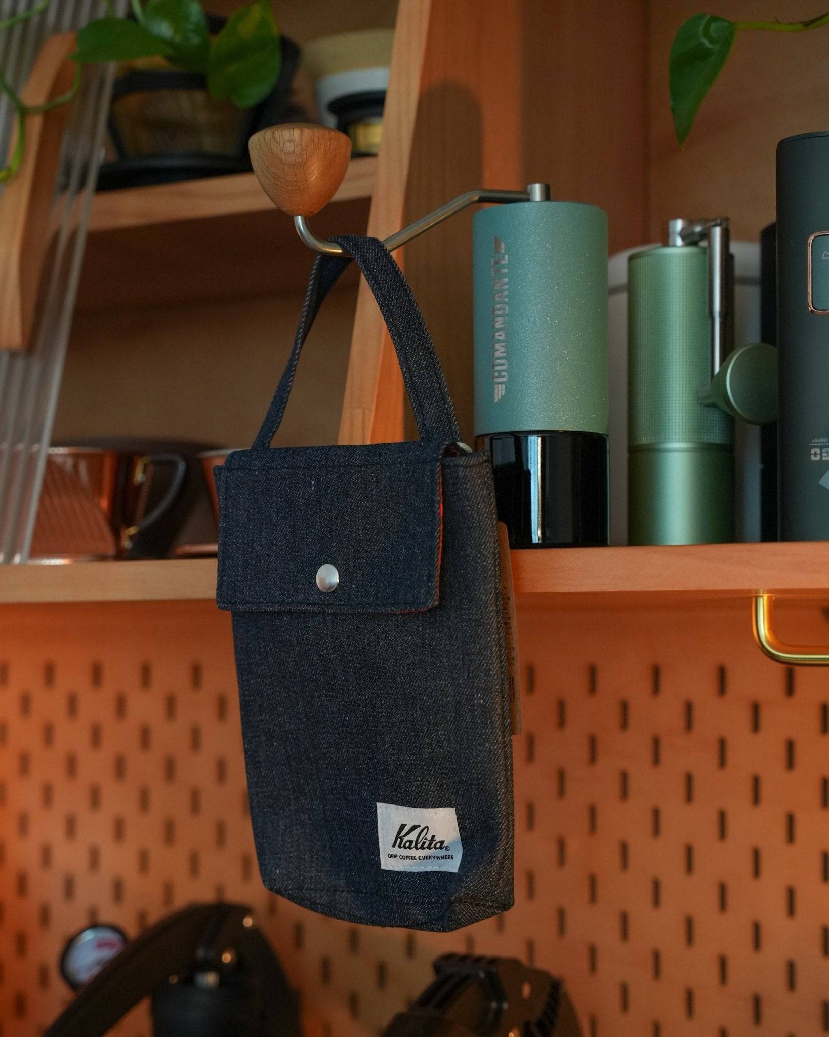 Stylish Kalita denim coffee filter storage bag hanging on wooden shelf, surrounded by coffee accessories and green plants
