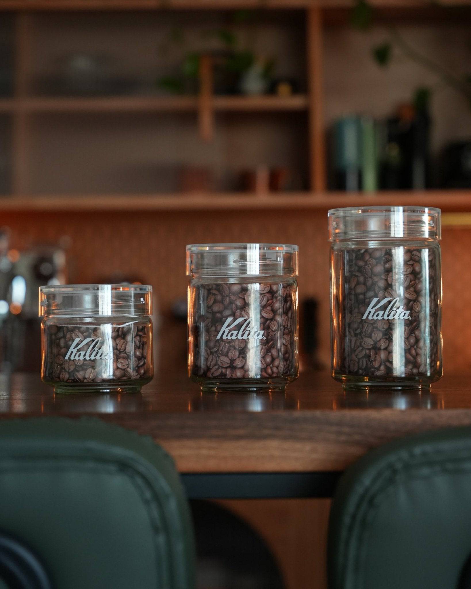 Three transparent Kalita glass coffee storage jars of different sizes filled with coffee beans, displayed on a wooden counter in a cozy cafe setting