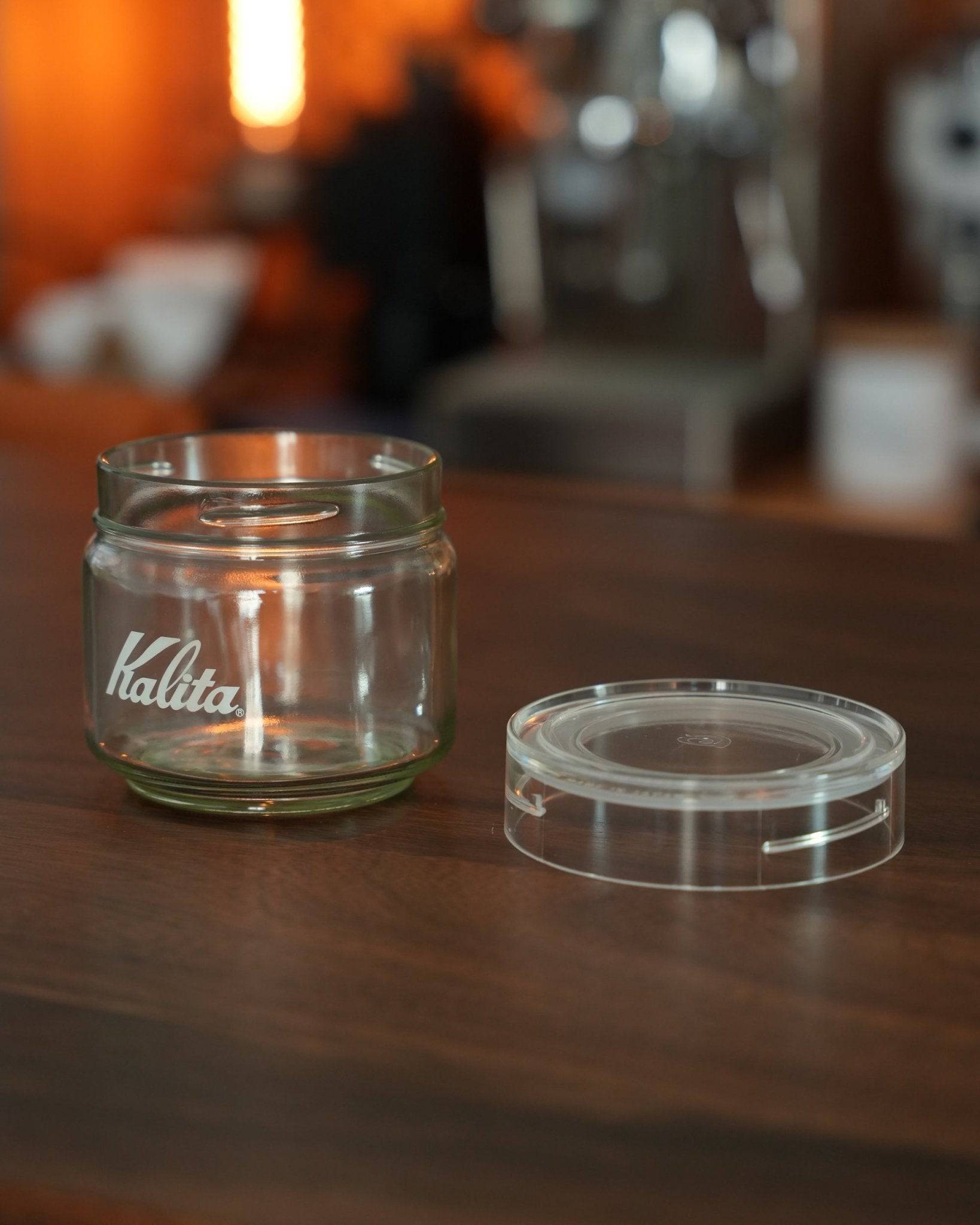 Kalita glass coffee storage jar with transparent lid on wooden surface, warm ambient lighting, showcasing sleek design and brand logo