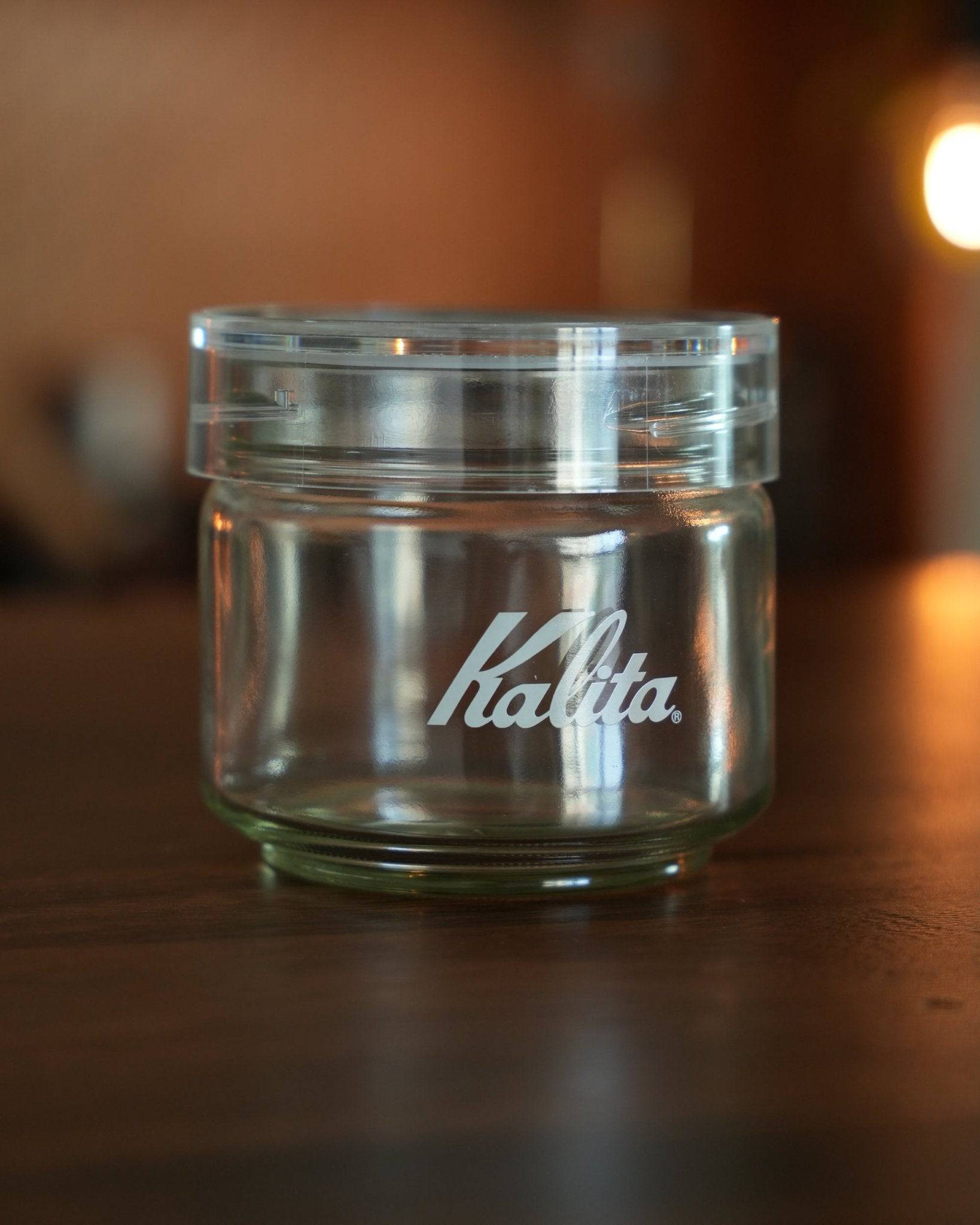 Kalita glass coffee storage jar with airtight lid, transparent container with white logo, placed on wooden surface in warm lighting