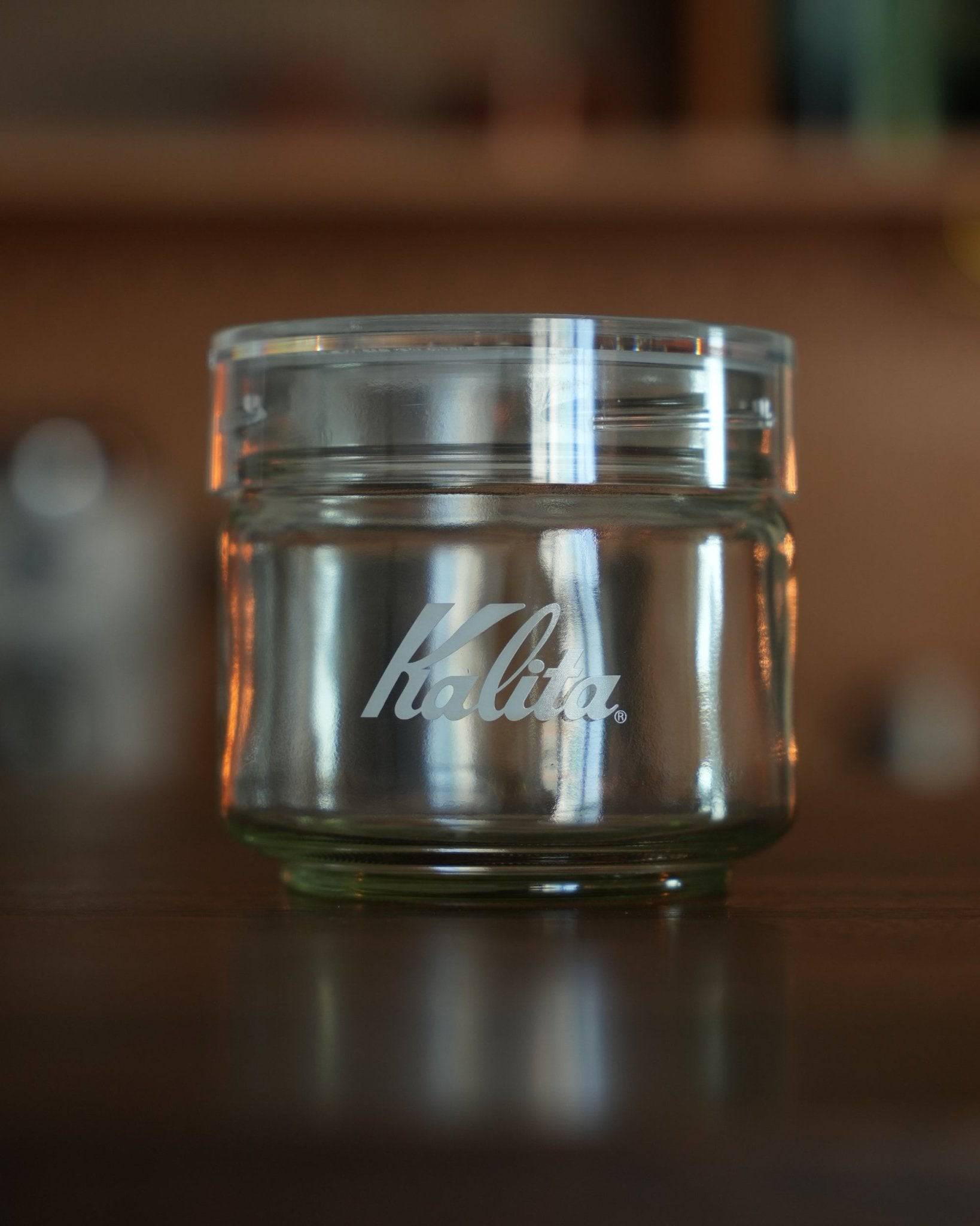 Kalita glass coffee storage jar with airtight lid, 150g capacity, transparent design showcasing Kalita logo, ideal for preserving coffee beans freshness, displayed on wooden surface