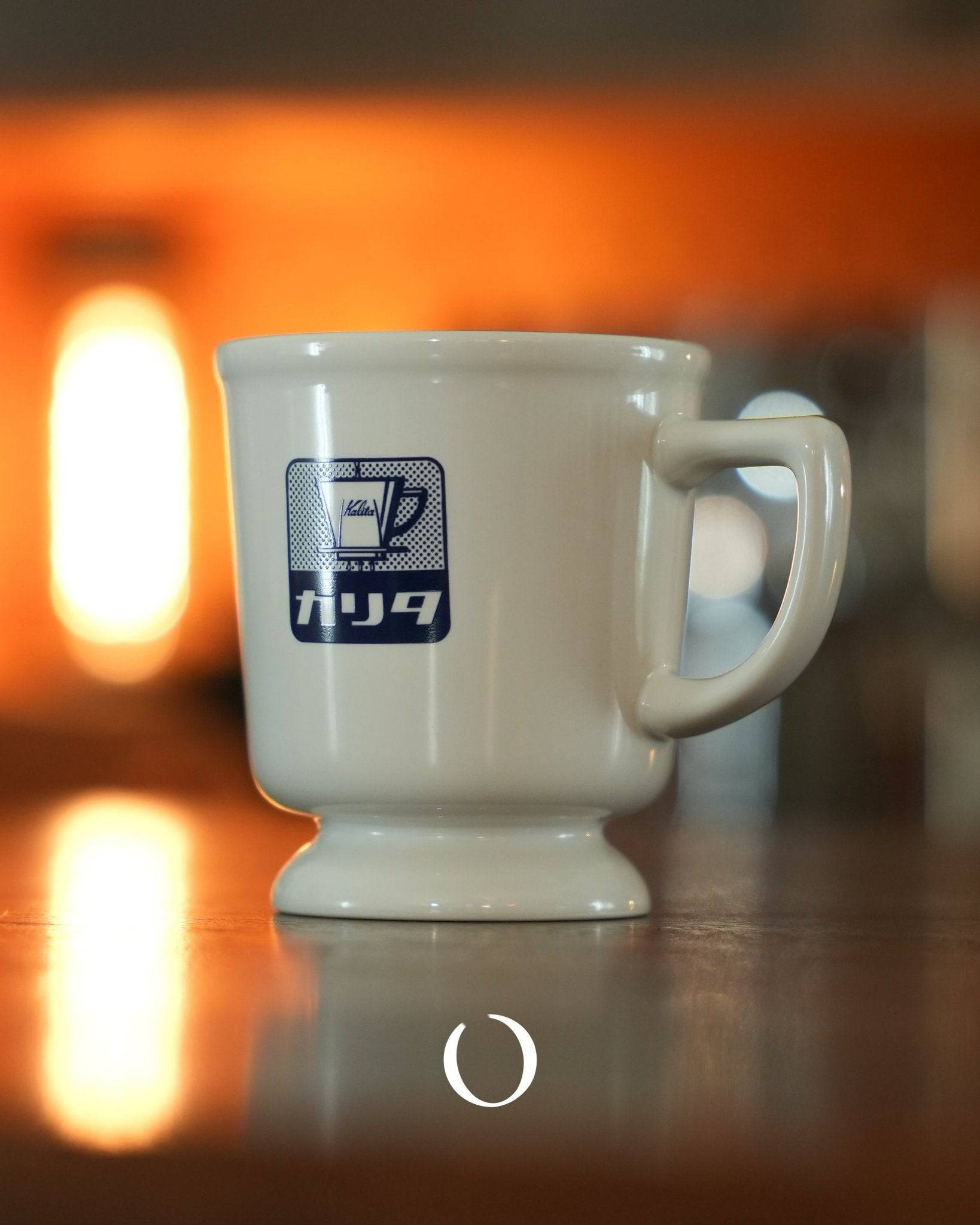 Vintage Kalita coffee mug with blue logo, white ceramic, retro design, on reflective surface with warm orange background lighting, showcasing Showa era Japanese aesthetic