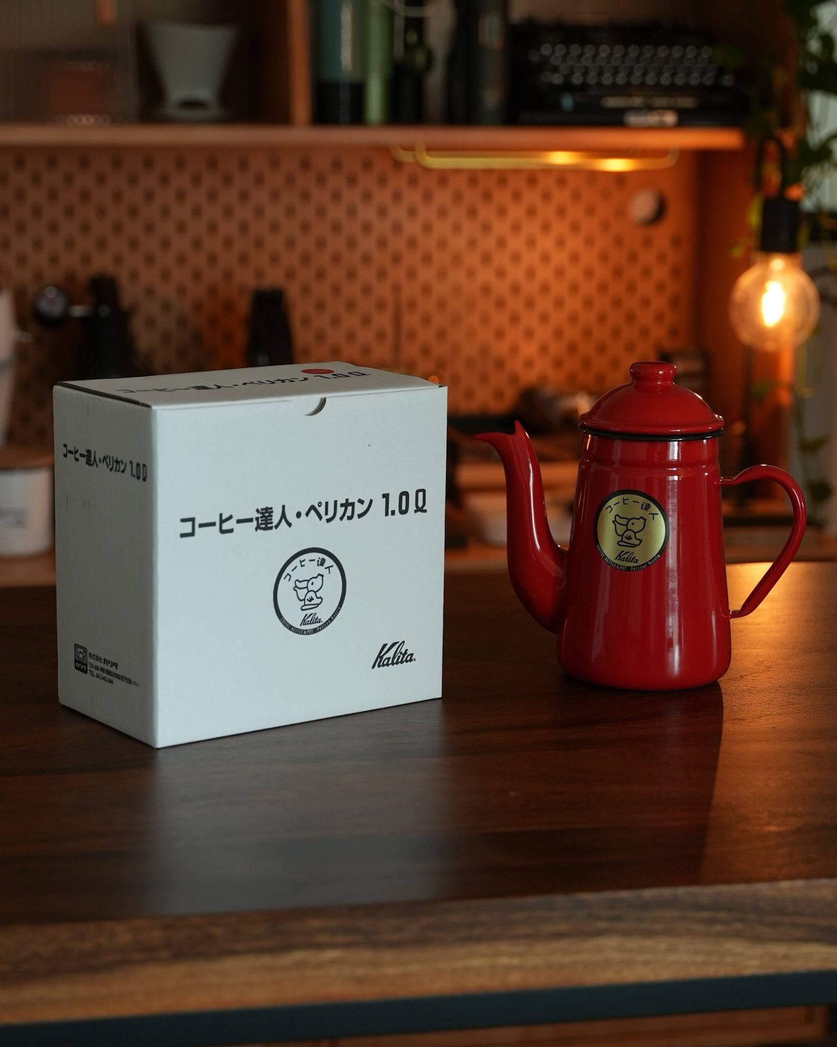 Red Kalita enamel pour-over kettle with gooseneck spout next to its product box on wooden counter, warm lighting, cozy coffee brewing setup