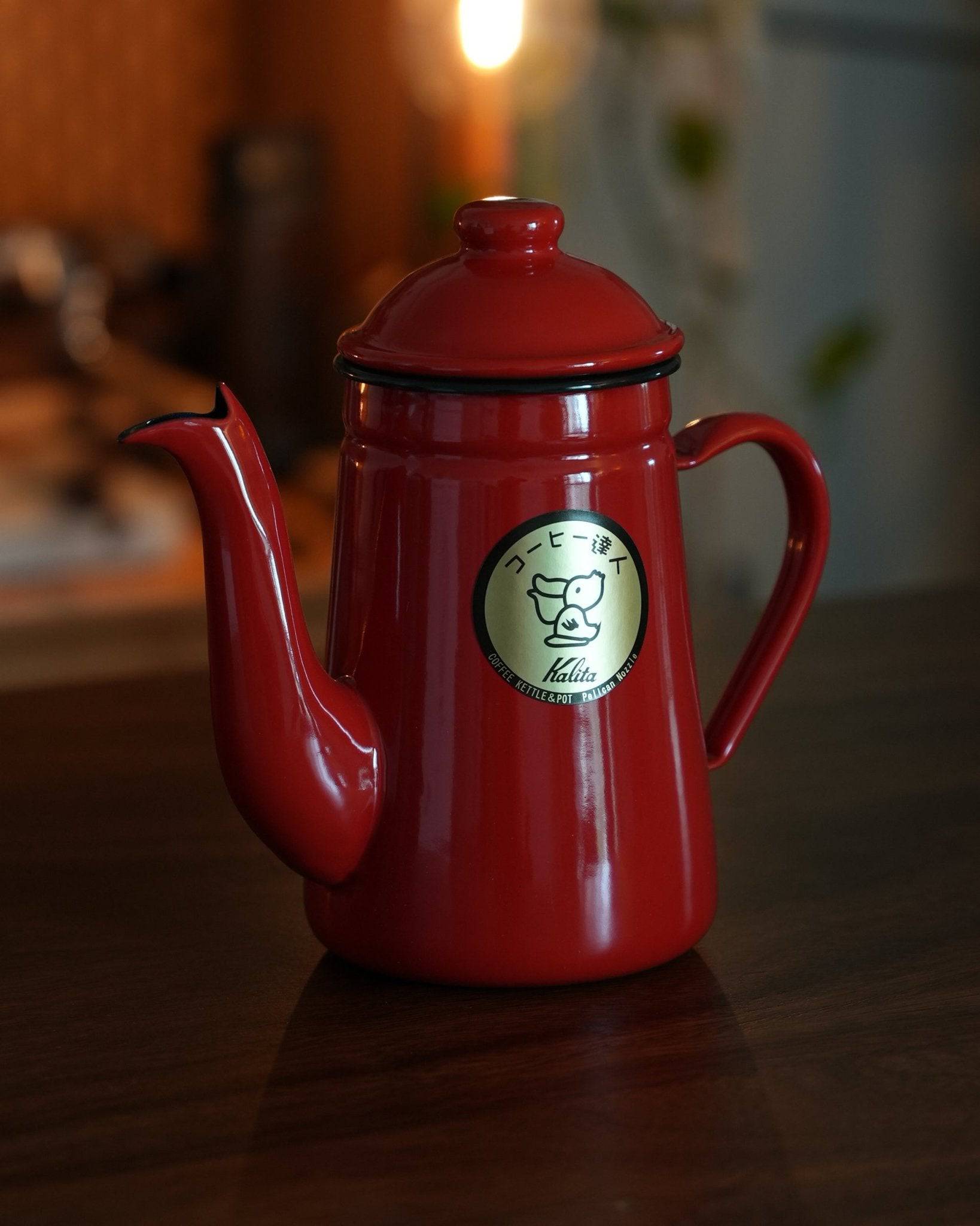 Shiny red Kalita enamel pour-over kettle with curved spout and handle, featuring brand logo, positioned on dark surface in warm lighting, ideal for precise coffee brewing
