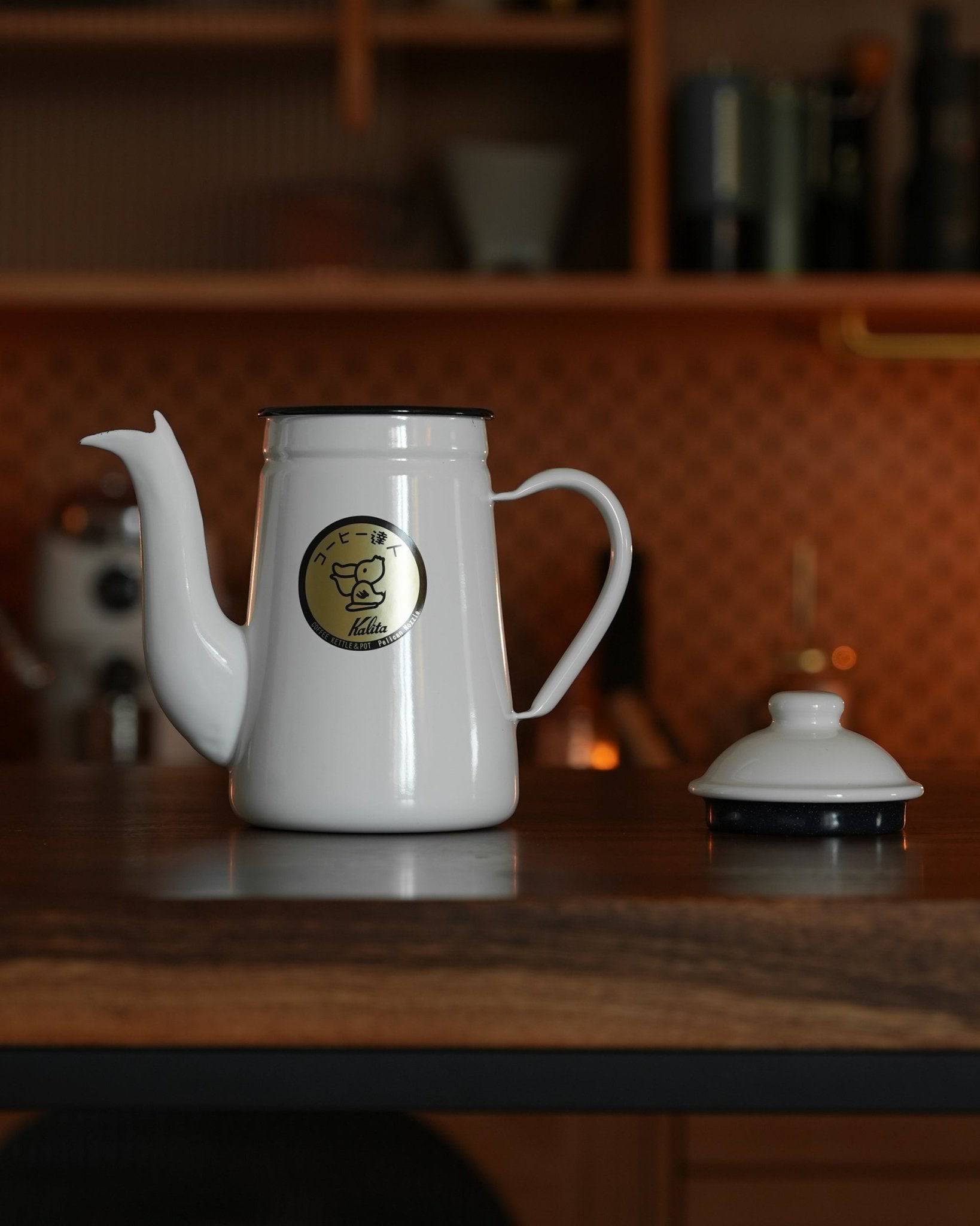 White Kalita enamel kettle with goose-neck spout on kitchen counter, featuring gold logo and detached lid, perfect for precision pour-over coffee brewing