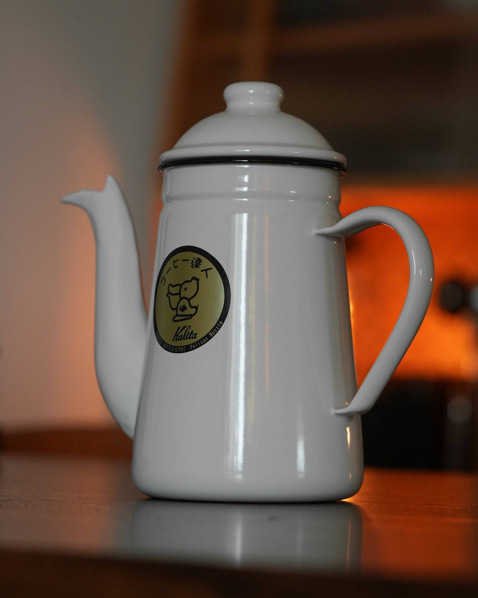 Kalita white enamel pour-over kettle with gooseneck spout and brand logo on wooden surface, orange glow in background