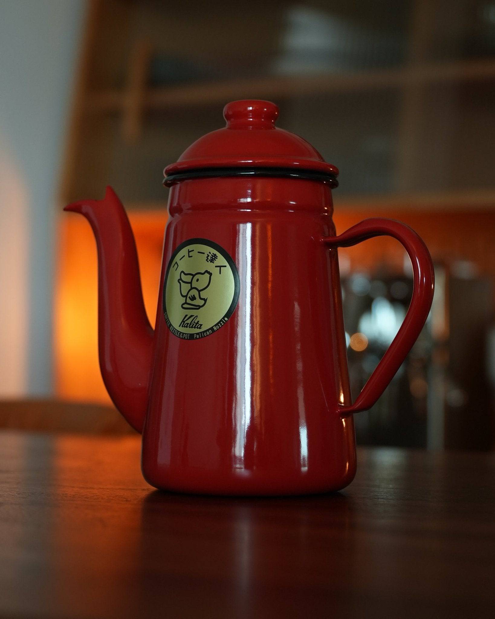 Vibrant red Kalita enamel pour-over kettle with crane-neck spout, 1L capacity, featuring brand logo on wooden table with warm ambient lighting