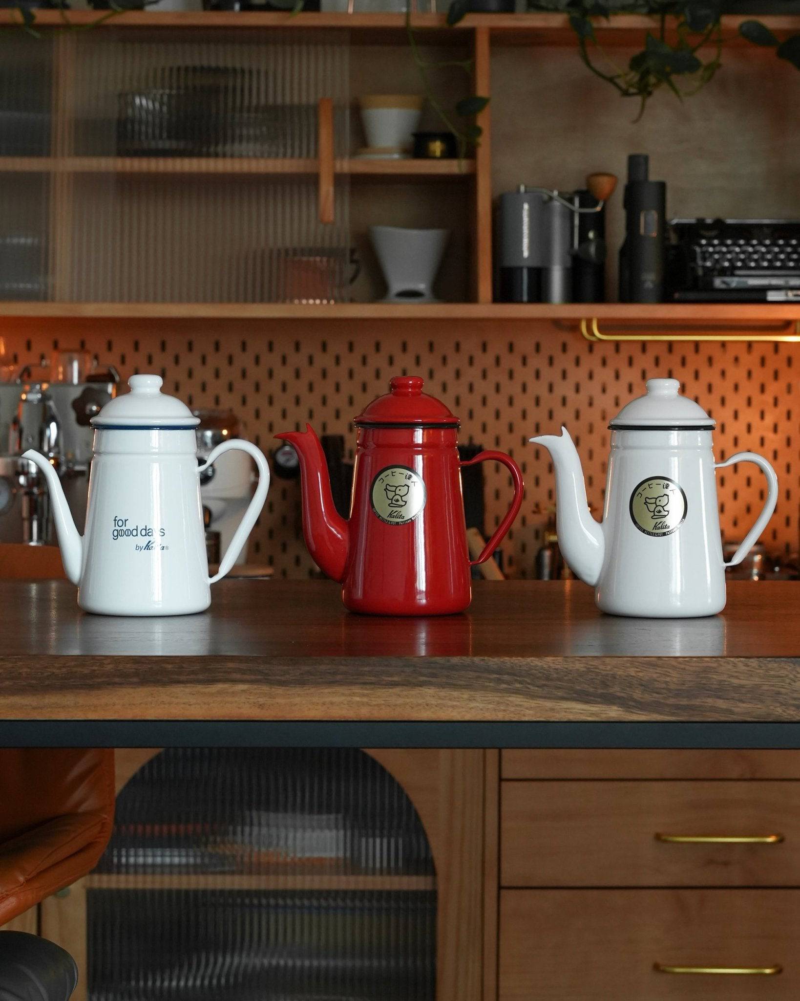 Three Kalita enamel coffee kettles in white, red, and light blue colors displayed on a kitchen counter with wooden shelves and appliances in the background