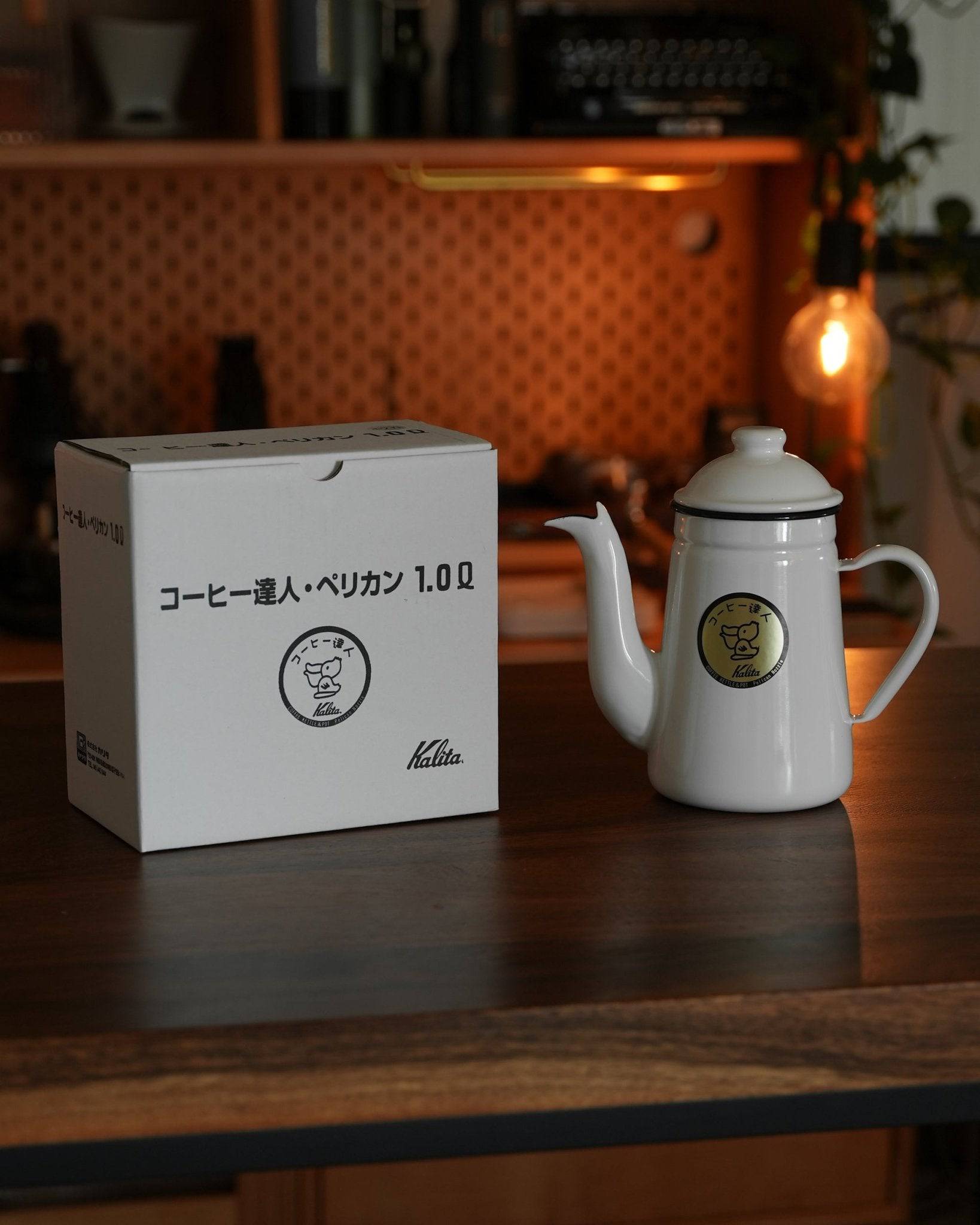 White Kalita enamel kettle with gooseneck spout and product box on wooden counter, warm lighting, coffee brewing equipment in background