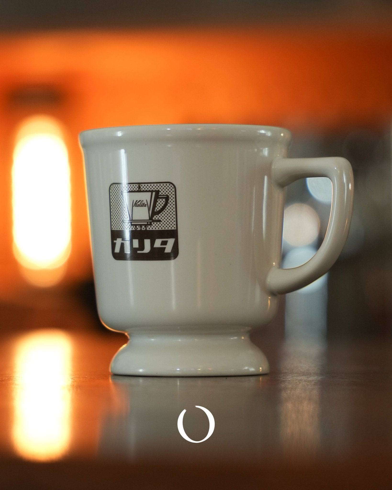 Kalita Showa era retro coffee mug in white ceramic with vintage logo, photographed against warm orange background, reflecting nostalgic Japanese cafe ambiance