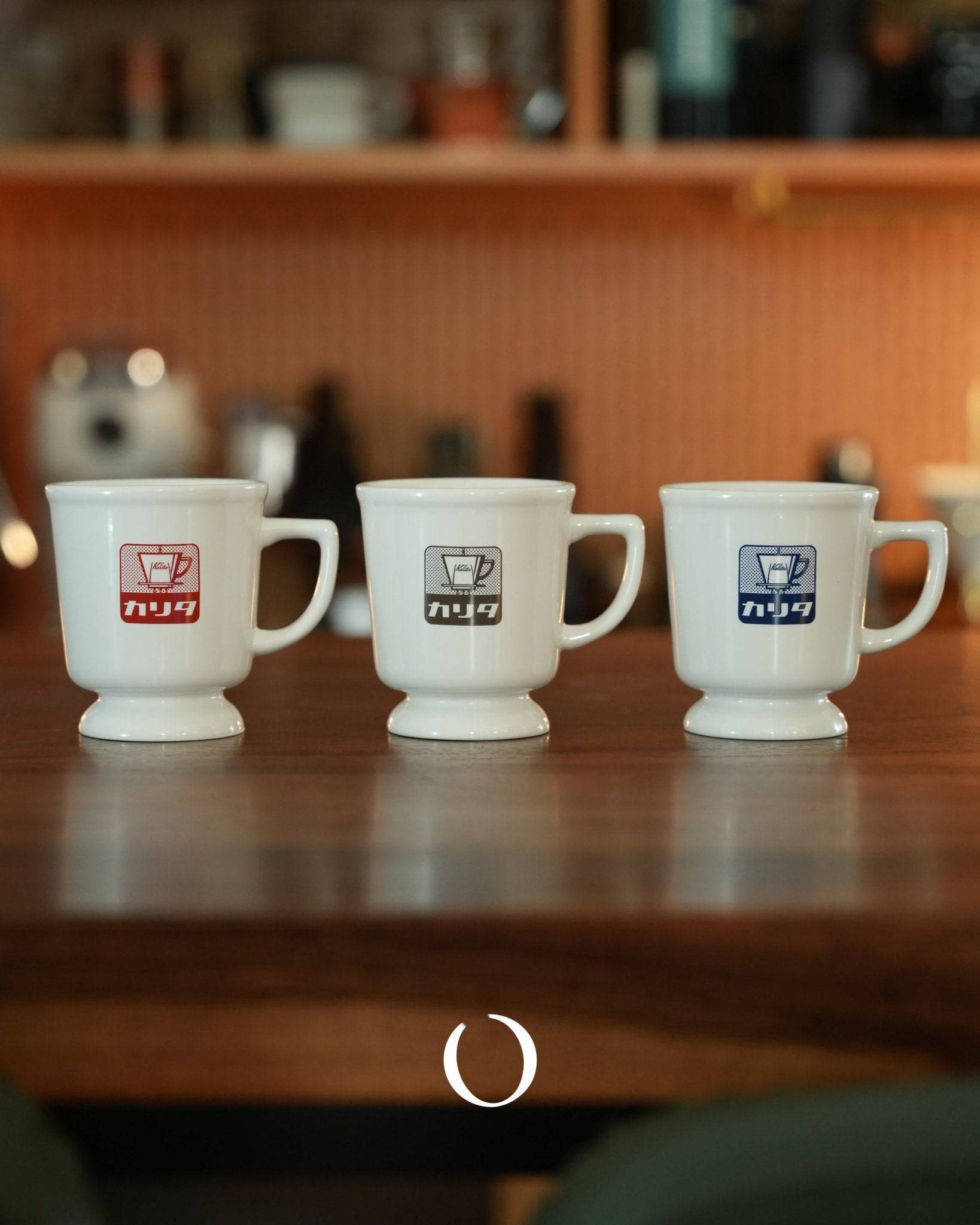 Three vintage Kalita coffee mugs in white with red, white, and blue logos, lined up on a wooden surface with a cozy kitchen background, showcasing retro Showa era design and nostalgic cafe atmosphere