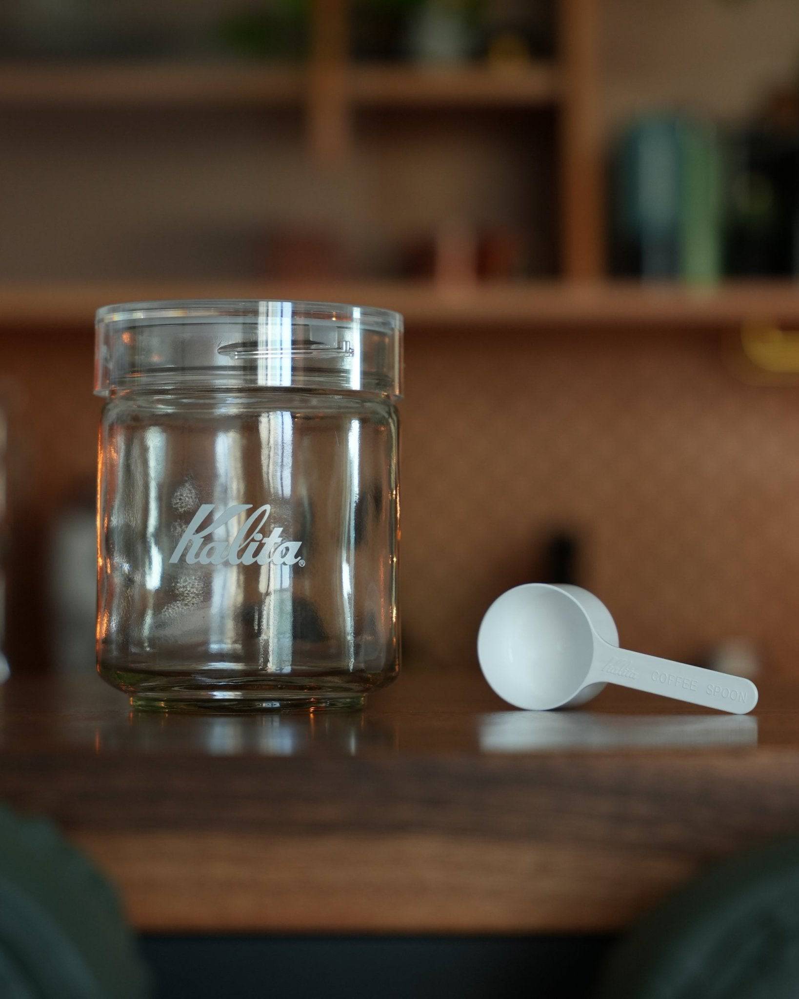 Kalita glass airtight coffee canister with Kalita logo, 250g capacity, transparent jar with white lid, accompanied by a white plastic measuring scoop, displayed on wooden surface