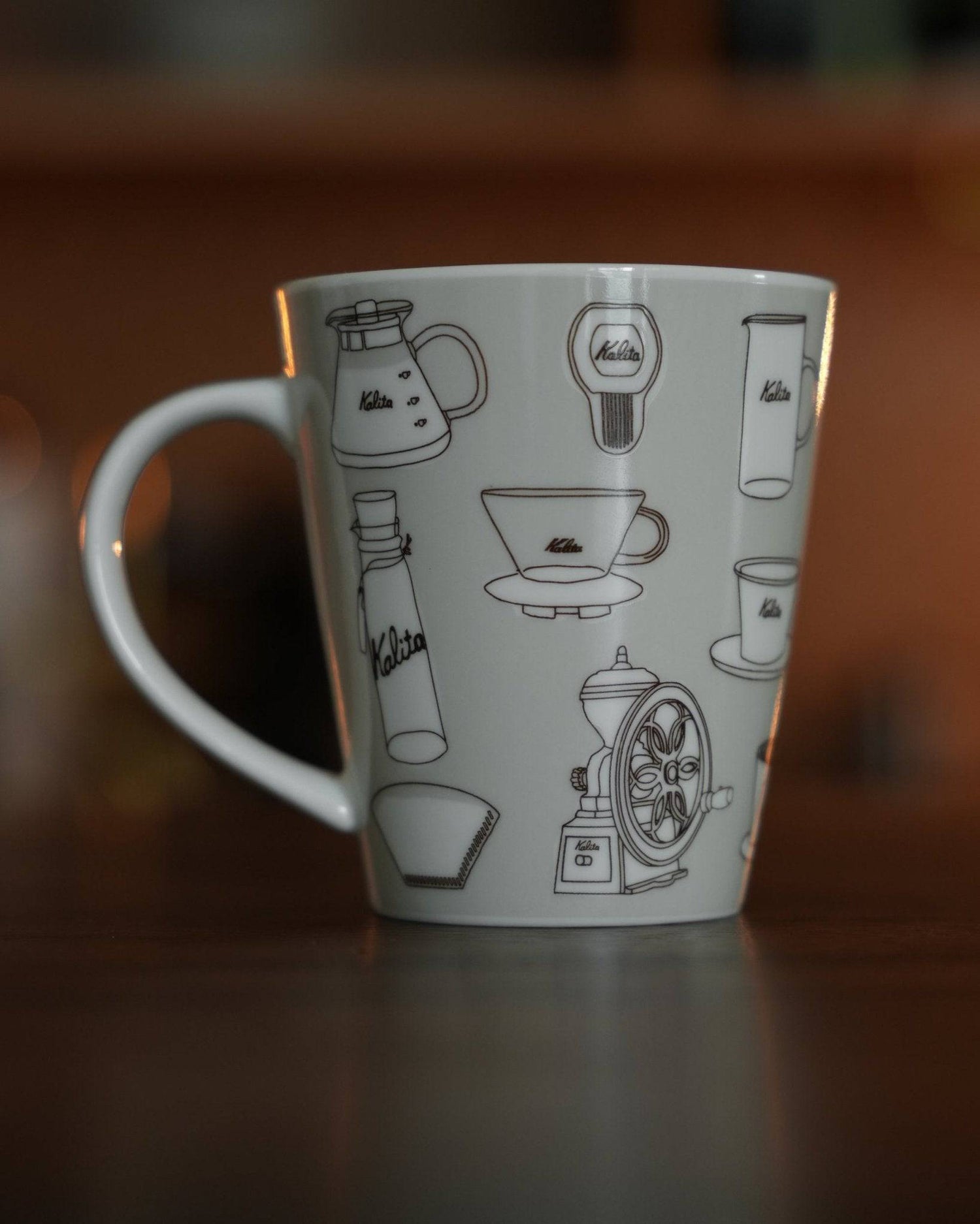 Kalita ceramic mug featuring illustrated coffee equipment designs on warm gray background, showcasing coffee pots, grinders, filters, and cups in black line art style, perfect for coffee enthusiasts and collectors