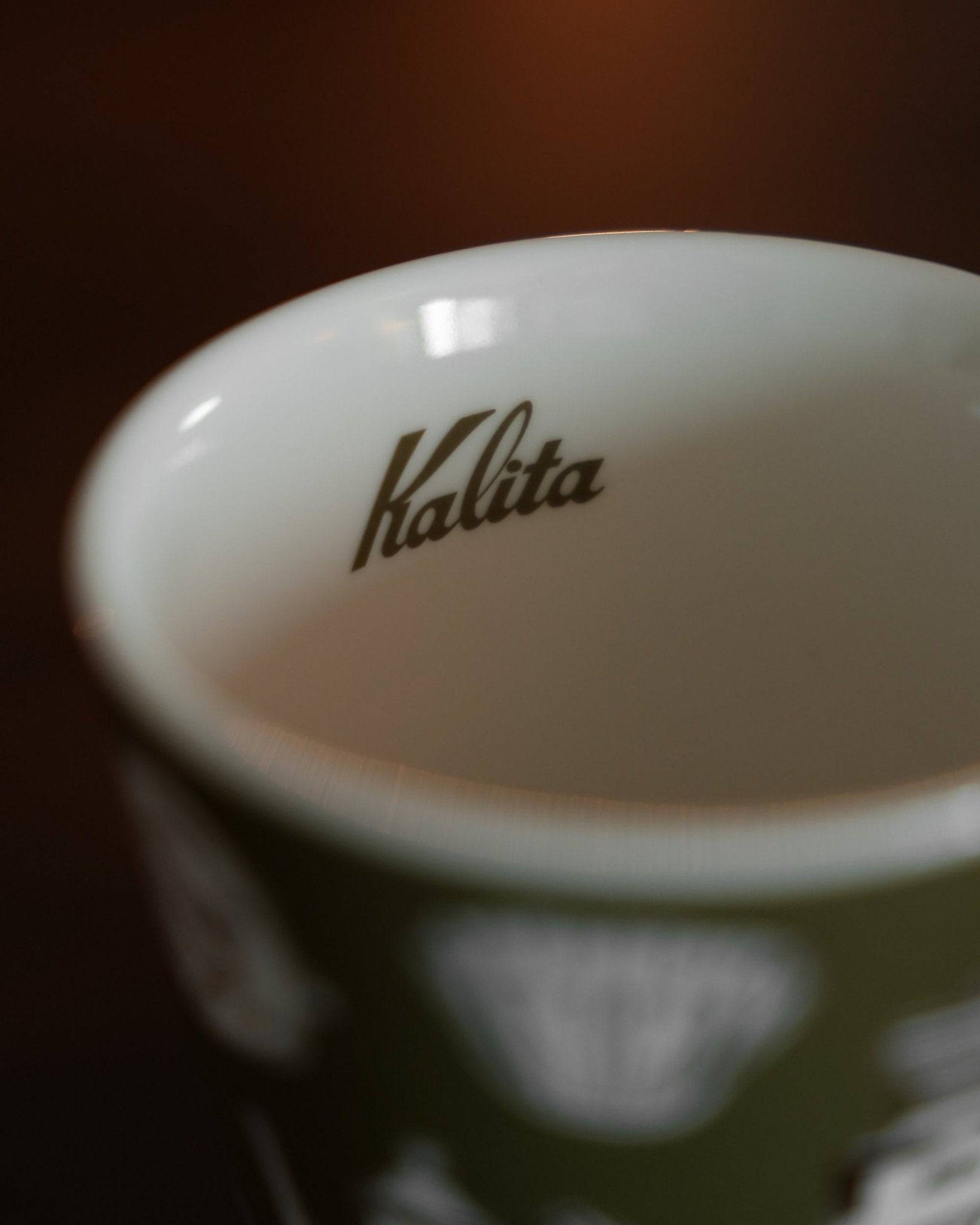 Close-up of a Kalita ceramic mug, showing the brand logo inside the cup. The exterior is dark green with white illustrations, contrasting with the white interior. The image highlights the quality and design of the Kalita coffee cup.