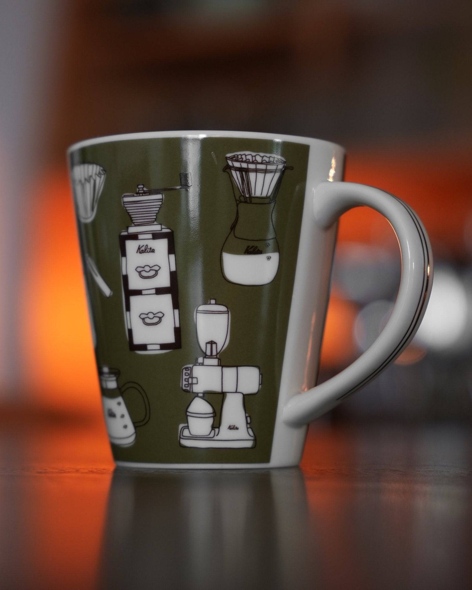 Kalita ceramic mug with khaki color featuring illustrated coffee equipment designs, including grinders, pour-over devices, and coffee makers, against a warm-toned background