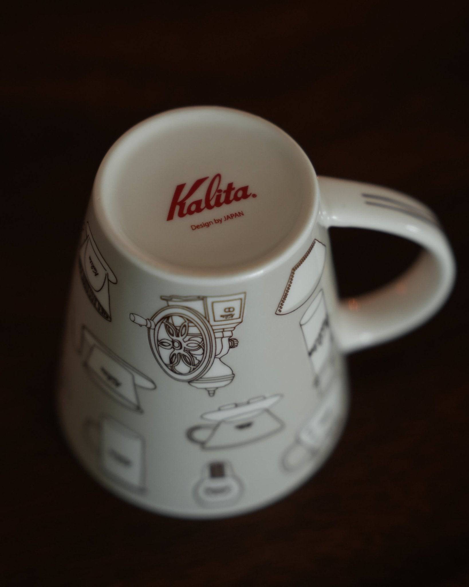Kalita ceramic mug in warm gray, featuring vintage coffee equipment designs and red logo, on dark wooden background. 300ml capacity, made in Japan.
