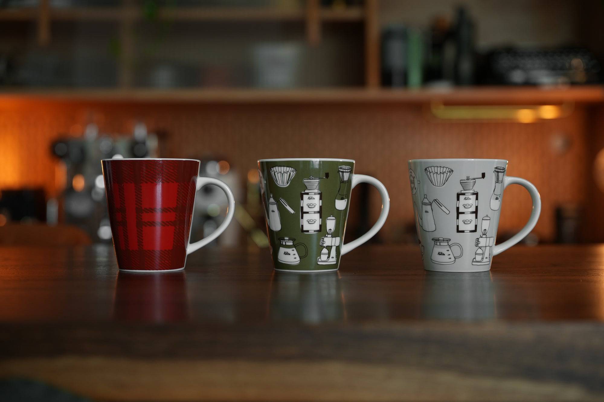 Three ceramic mugs on wooden surface: red plaid, green with coffee equipment illustrations, and white with coffee gear sketches. Kitchen background with warm lighting.
