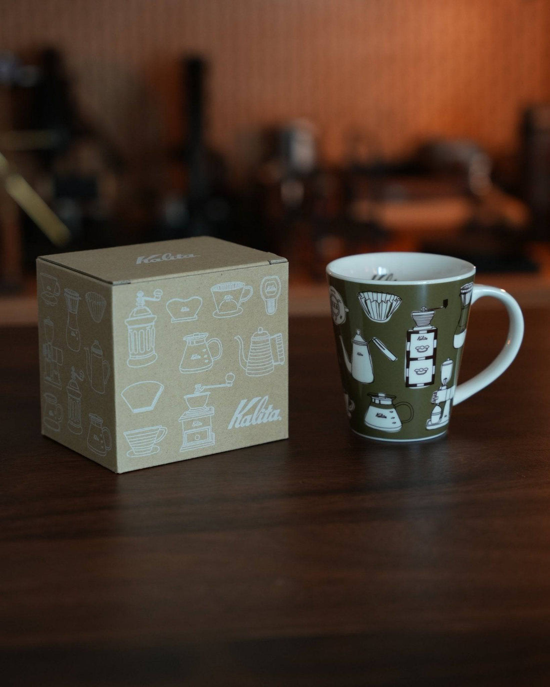 Kalita ceramic mug in khaki color with coffee equipment illustrations, displayed next to its matching box on wooden surface, coffee-making tools in background