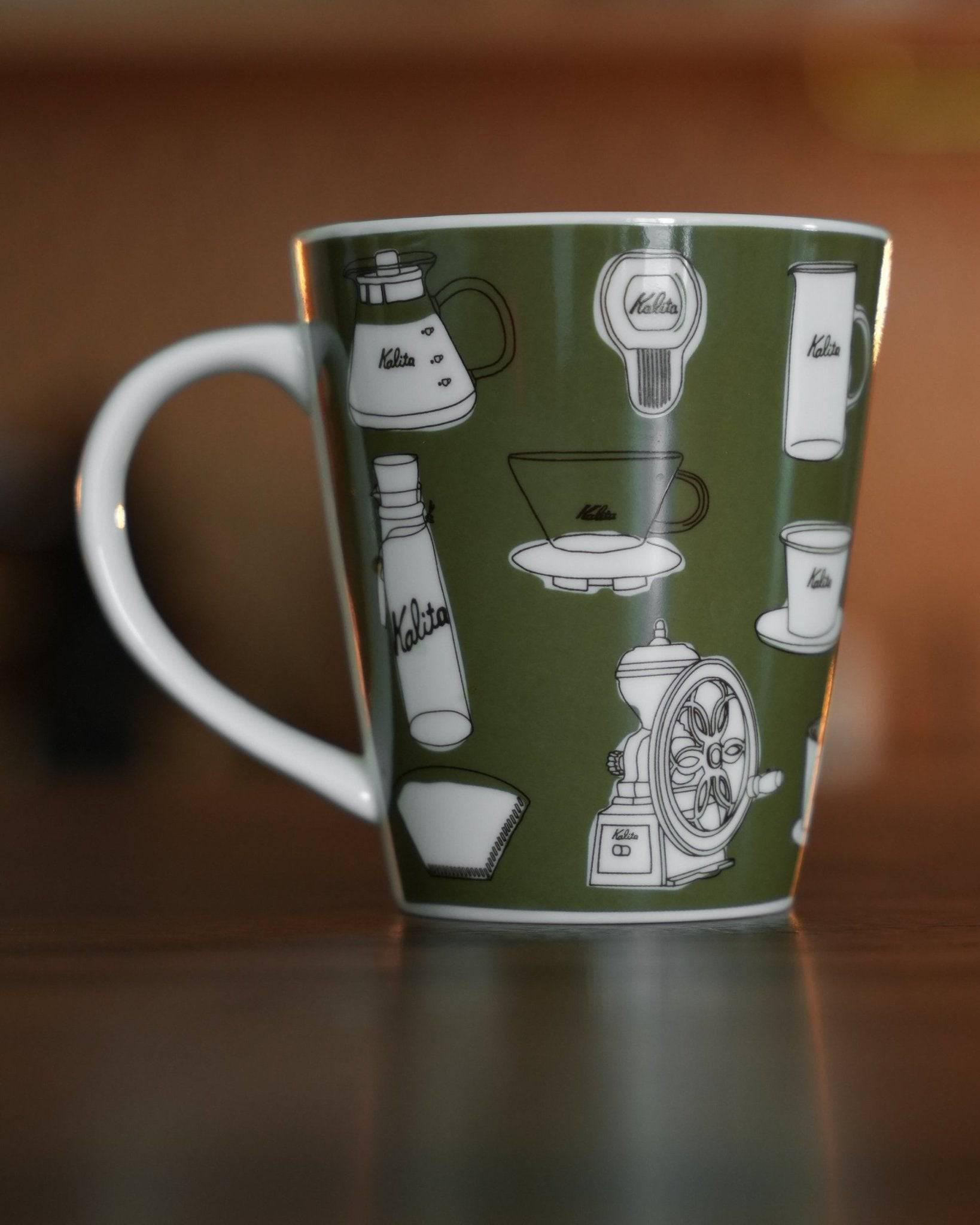 Kalita ceramic mug in khaki color featuring white illustrations of coffee equipment, including grinder, kettle, and filters, on a green background, showcasing brand&