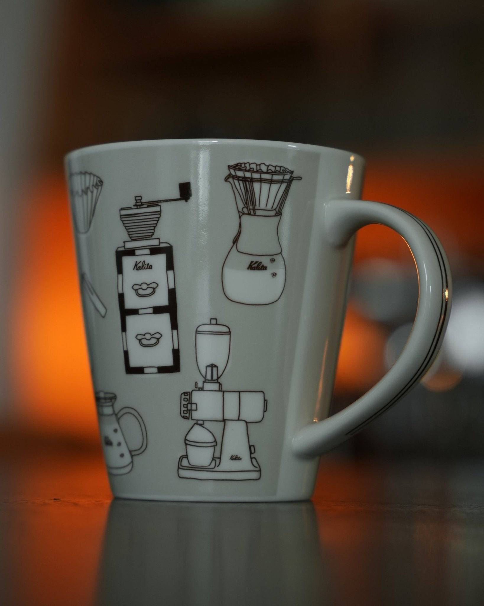 Kalita ceramic mug in warm gray, featuring black illustrations of classic coffee equipment including grinders, brewers, and filters, set against a warm-toned background
