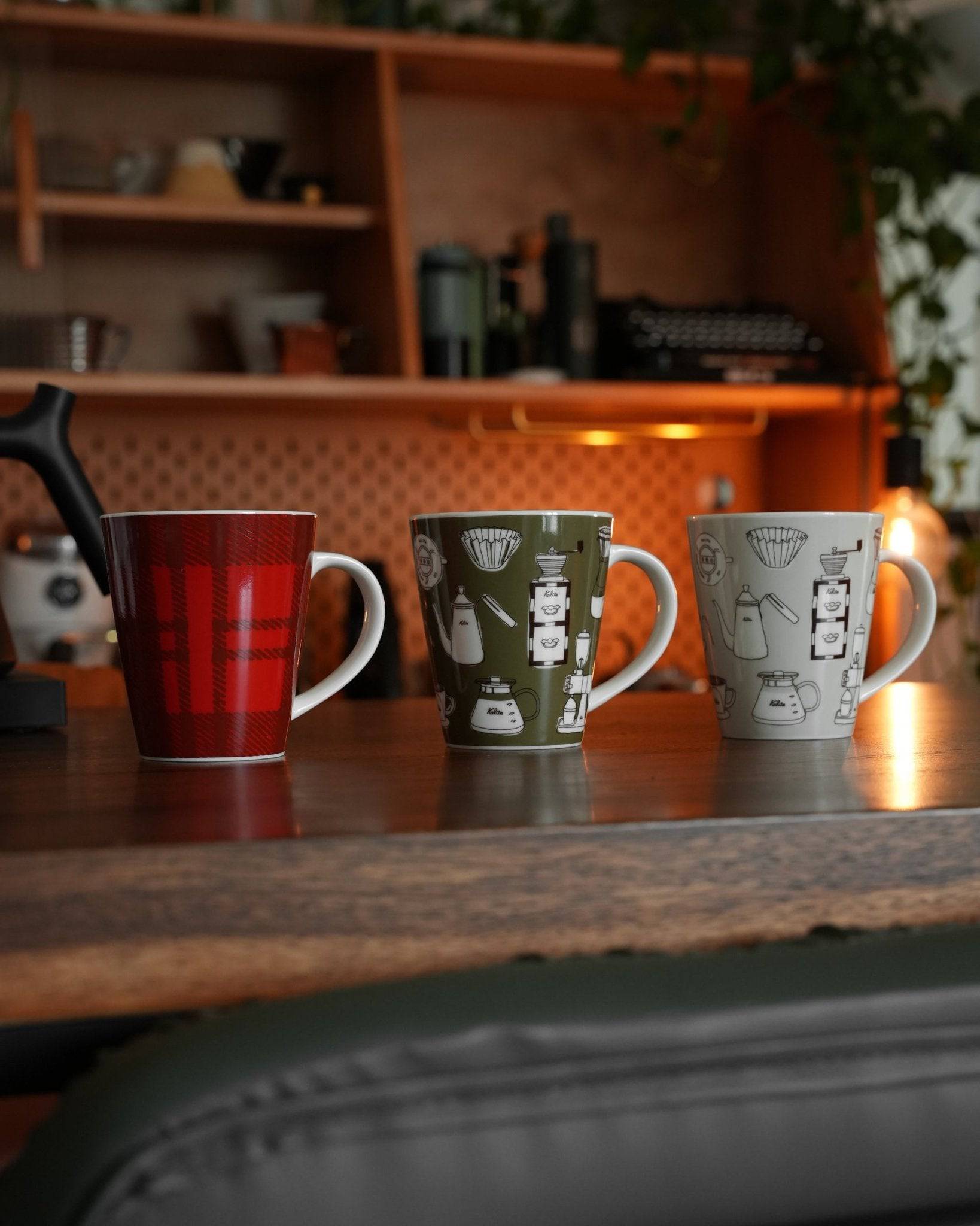 Three Kalita ceramic mugs on wooden counter, red checkered design, olive and white with coffee equipment illustrations. Warm kitchen ambiance with shelves in background.