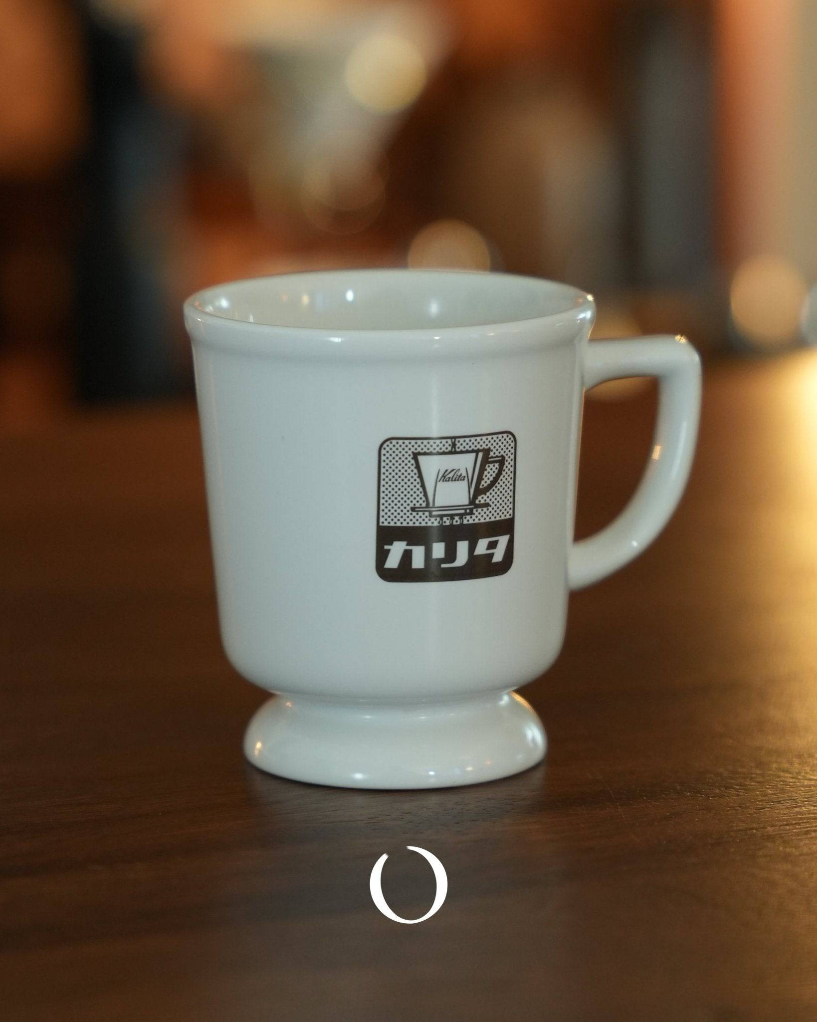 Vintage Kalita coffee mug with retro logo, white ceramic, classic design, wide rim, elevated base, on wooden surface with blurred background, nostalgic Japanese cafe atmosphere