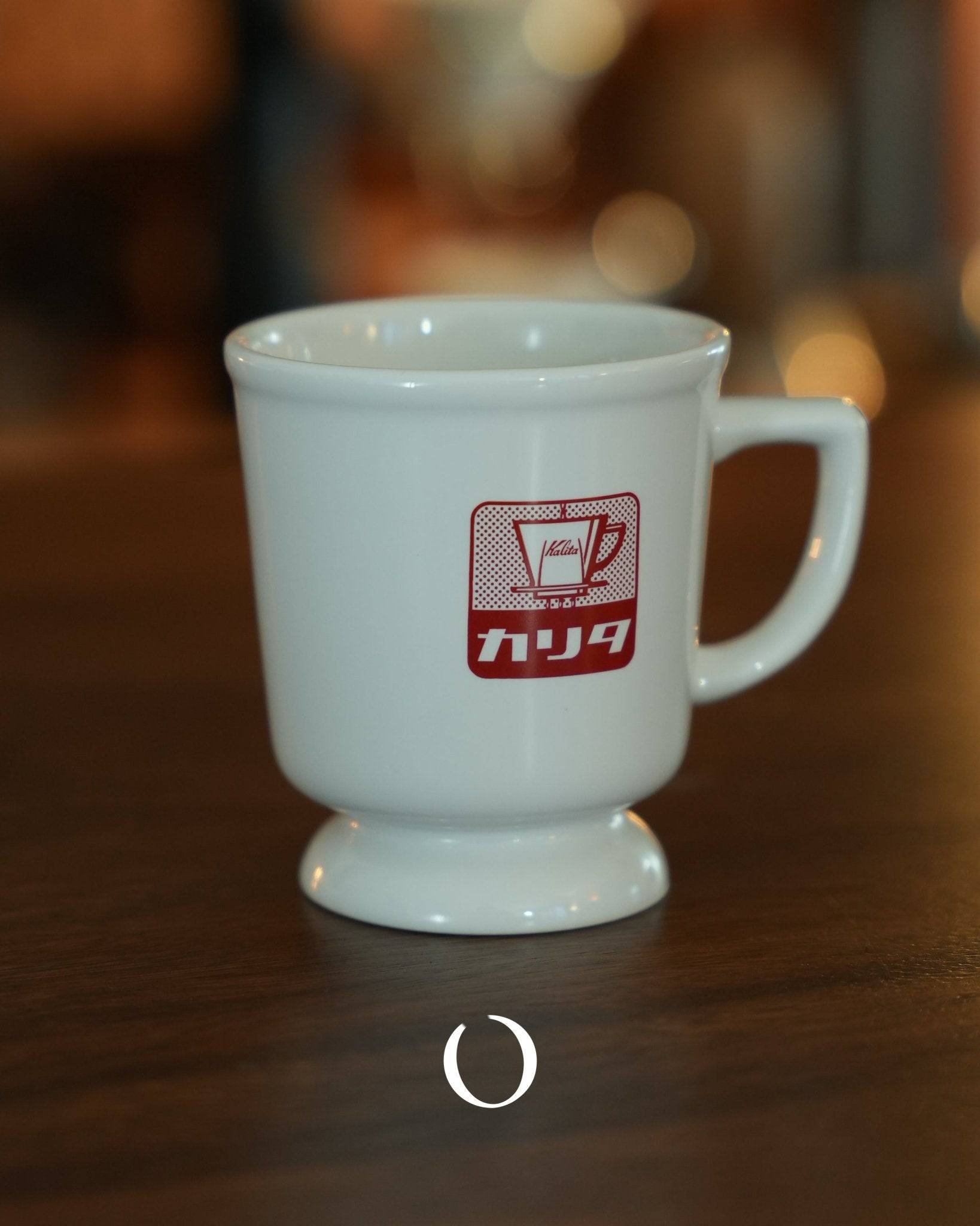Vintage Kalita coffee mug with red logo, white ceramic, retro Showa era design, wide mouth and elevated base, on wooden table with blurred background