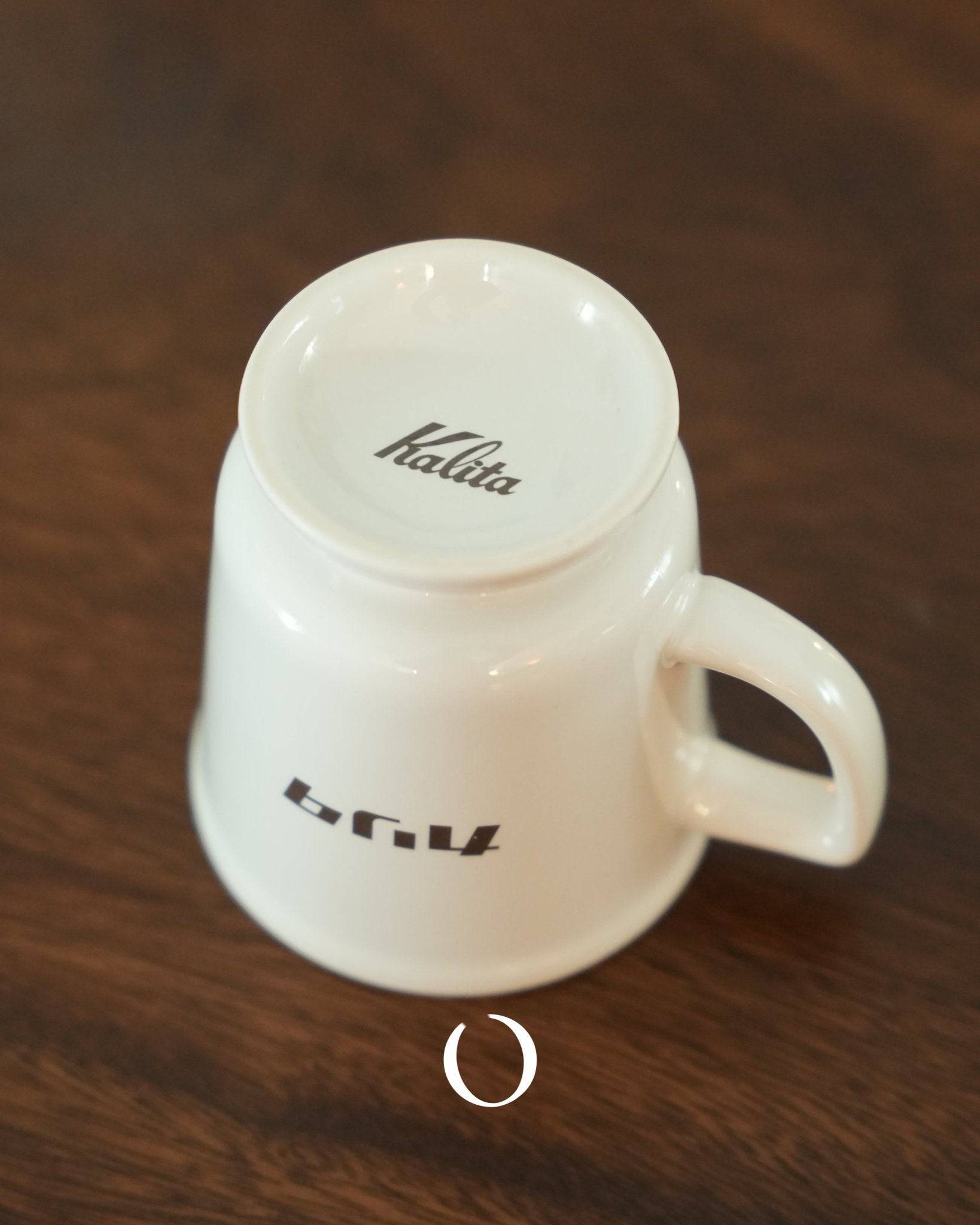 Kalita vintage-style ceramic coffee mug in white, featuring retro design with brand logo on bottom and side. Wide mouth, thick walls for heat retention, sitting on wooden surface. Classic Japanese Showa era aesthetic for nostalgic cafe experience.