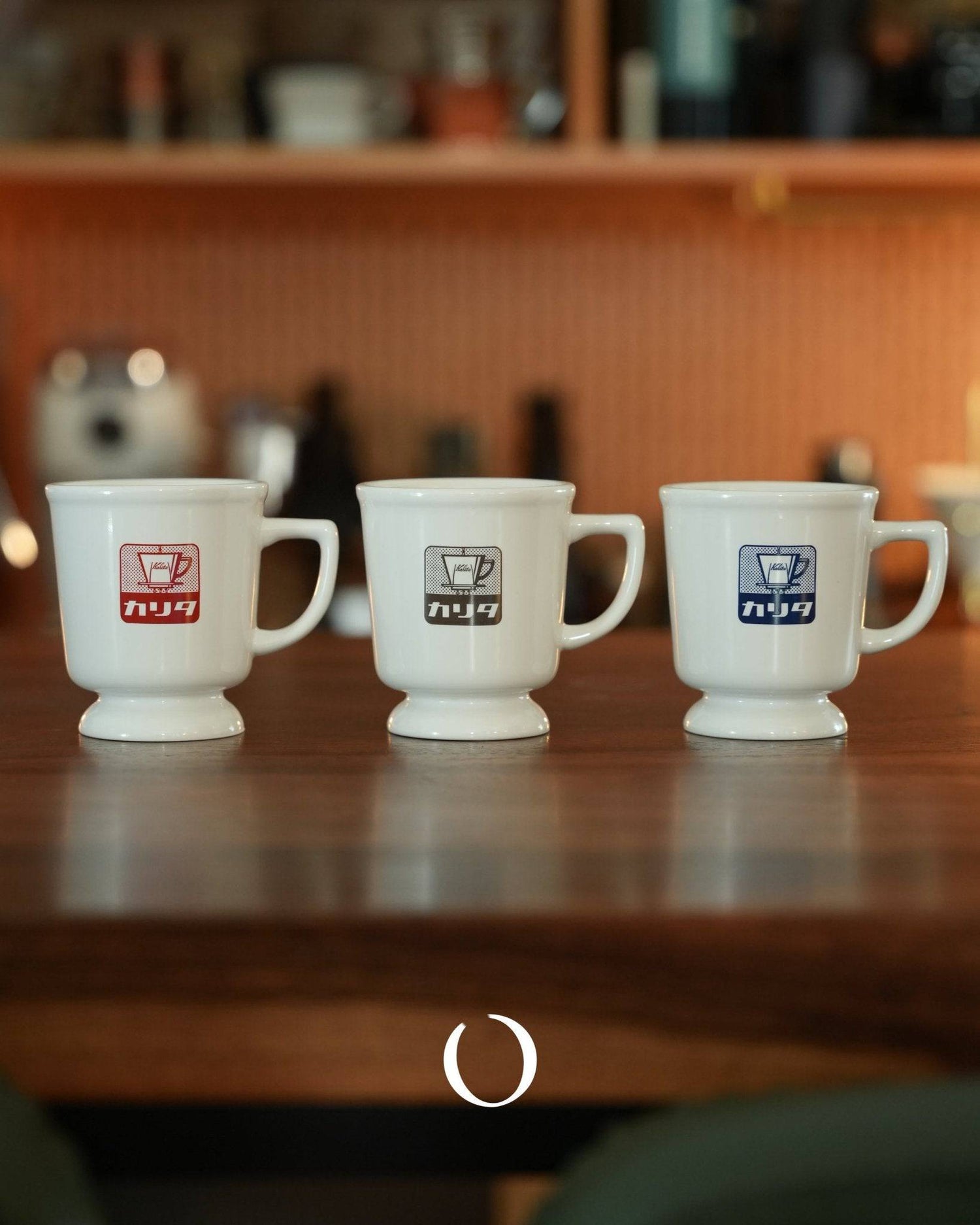 Three vintage Kalita coffee mugs in white ceramic with red, white, and blue logos, displayed on a wooden table against a warm-toned background with shelves of coffee accessories