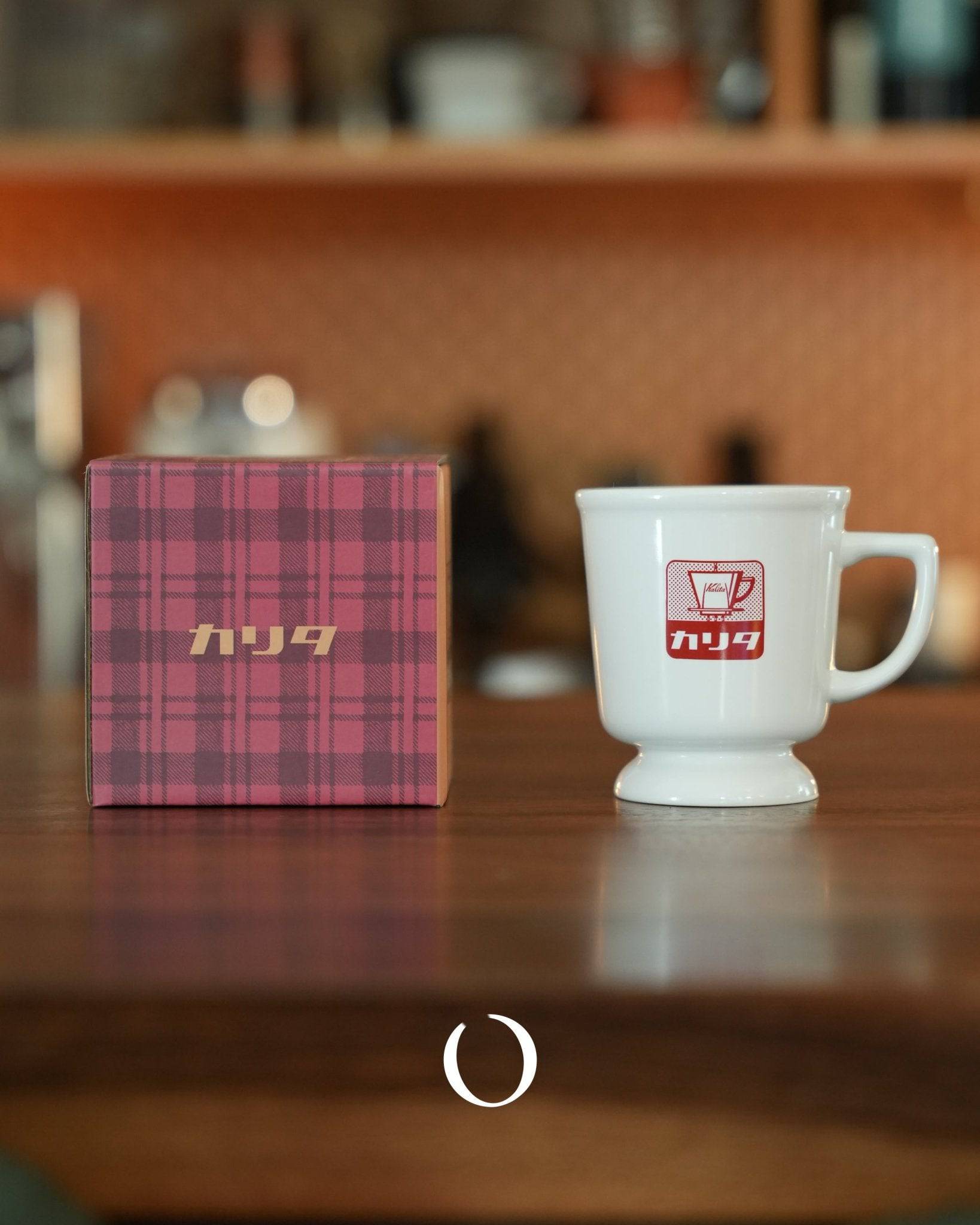 Vintage Kalita coffee mug with red logo next to plaid packaging box on wooden surface, retro Japanese coffee shop ambiance in background