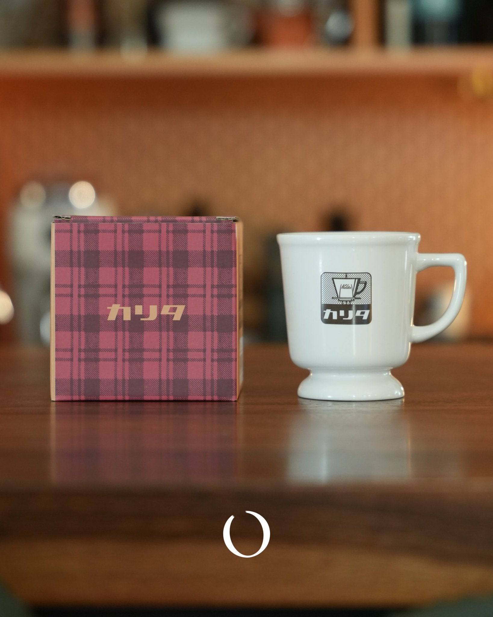 Vintage Kalita coffee mug in white ceramic with logo, displayed next to its plaid packaging box on wooden surface, warm retro ambiance