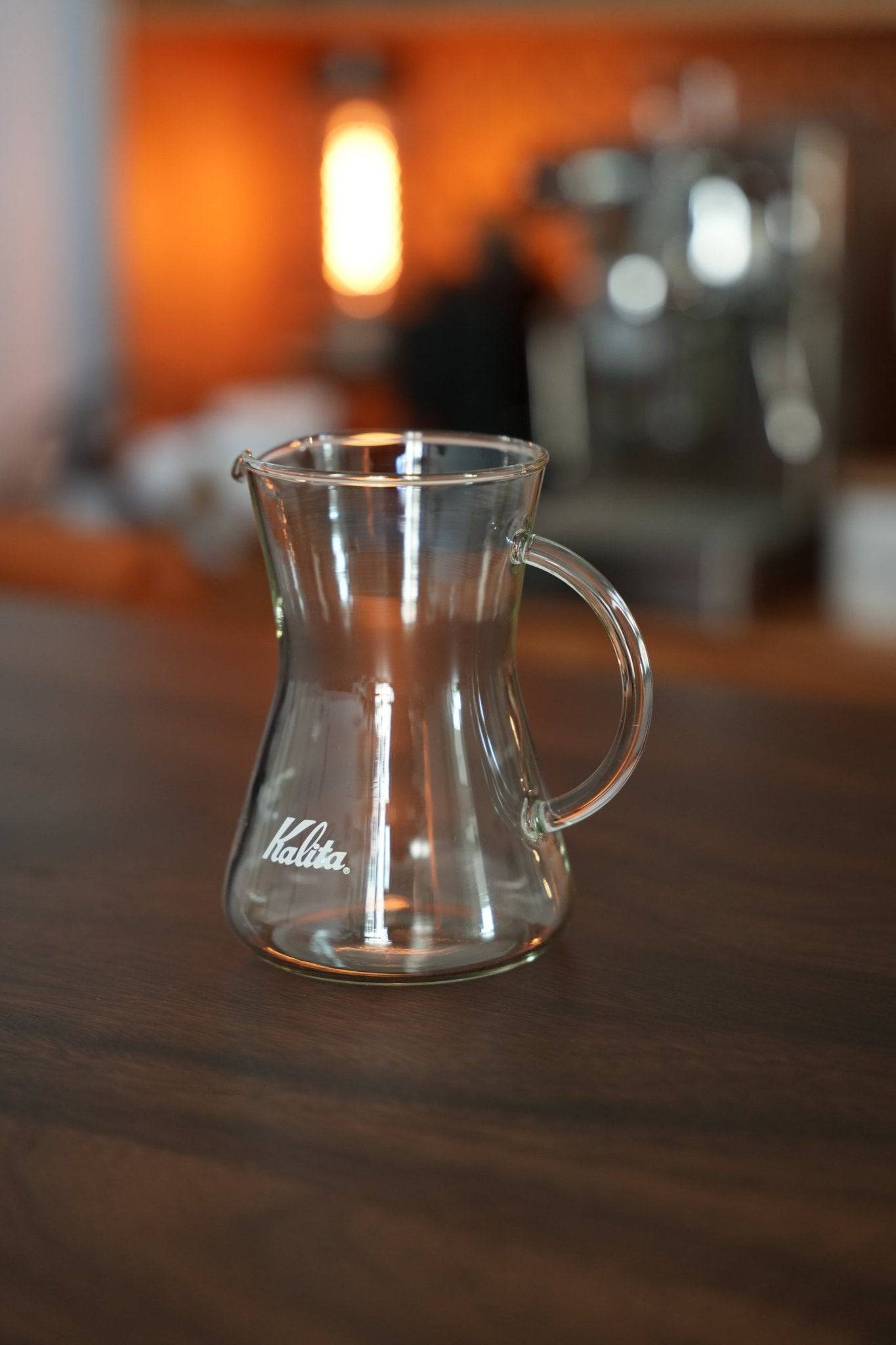 Kalita conical server 300 glass coffee carafe on wooden surface, showcasing its elegant design with Kalita logo. Warm ambient lighting highlights the transparent, heat-resistant glass structure. Coffee machine and light fixture visible in blurred background.
