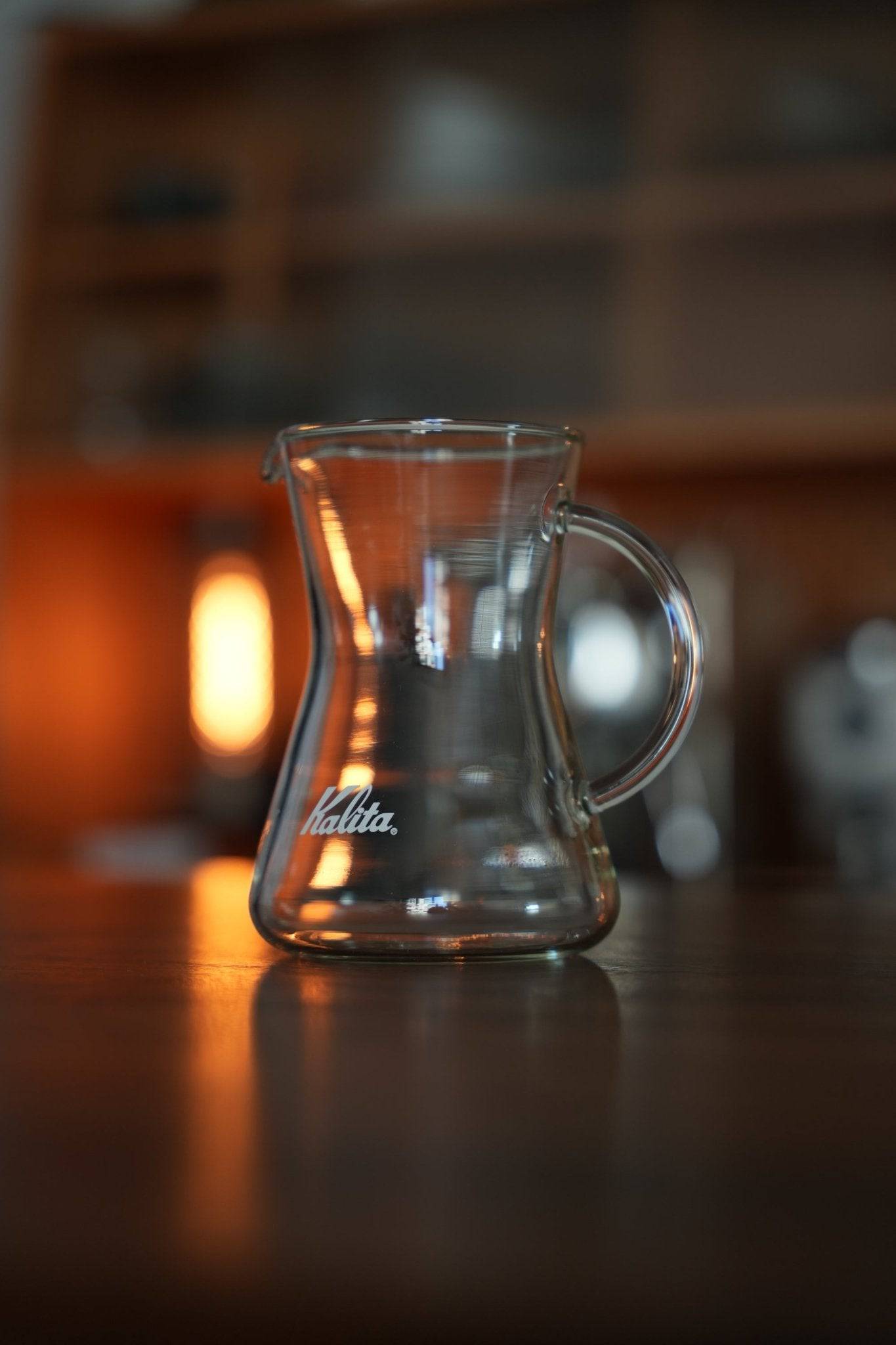 Kalita conical glass coffee server on wooden table with warm ambient lighting, showcasing its elegant design, heat-resistant properties, and versatility for various hot beverages