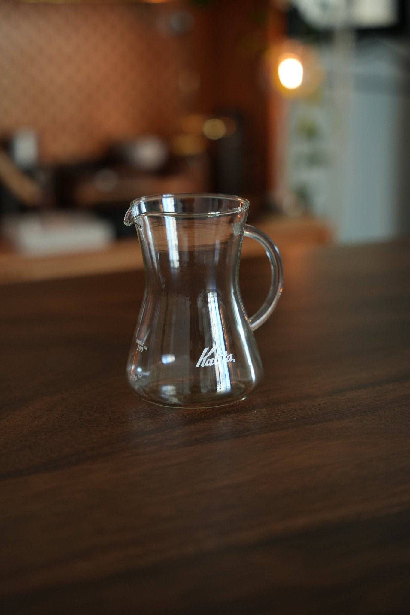 Kalita conical server 300 glass coffee carafe on wooden table in cozy kitchen, showcasing its elegant design, heat-resistant glass, and Kalita logo. Perfect for brewing and serving various hot beverages.