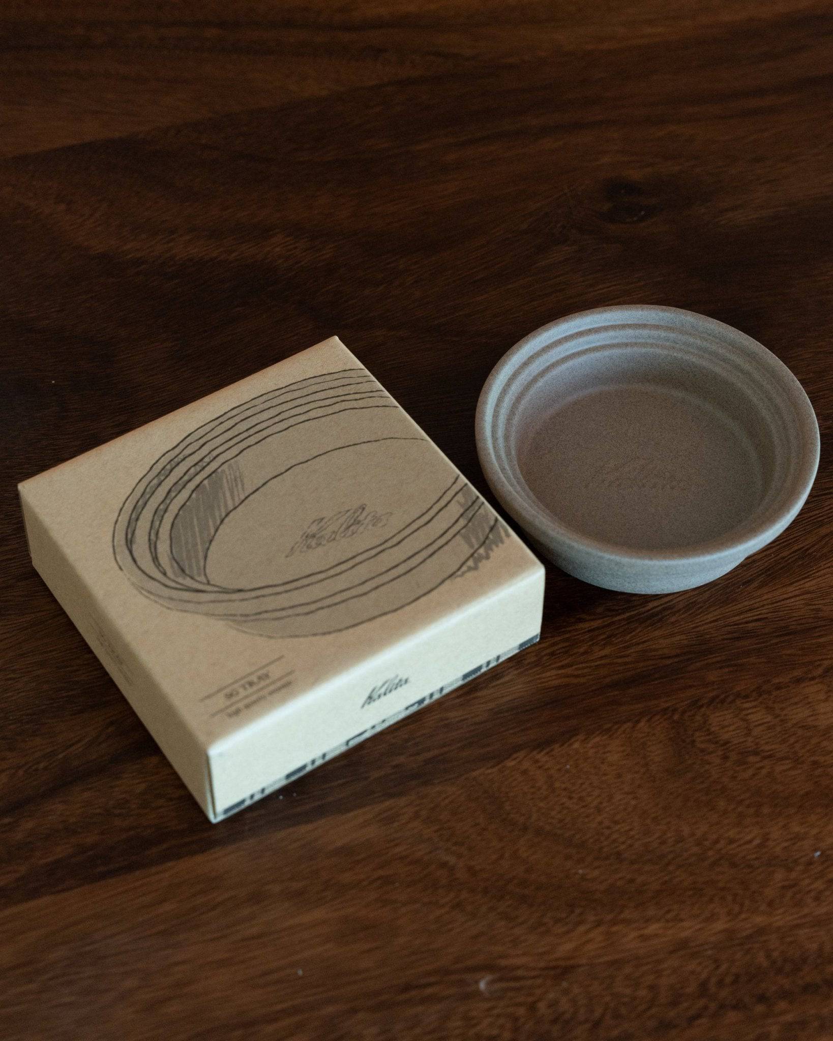 Kalita & HASAMI sandstone coffee dripper tray with box, showcasing Japanese Hasami-yaki craftsmanship. Circular tray with raised center ring, designed for Kalita wave drippers. Handcrafted ceramic in muted gray color, displayed on dark wood surface.
