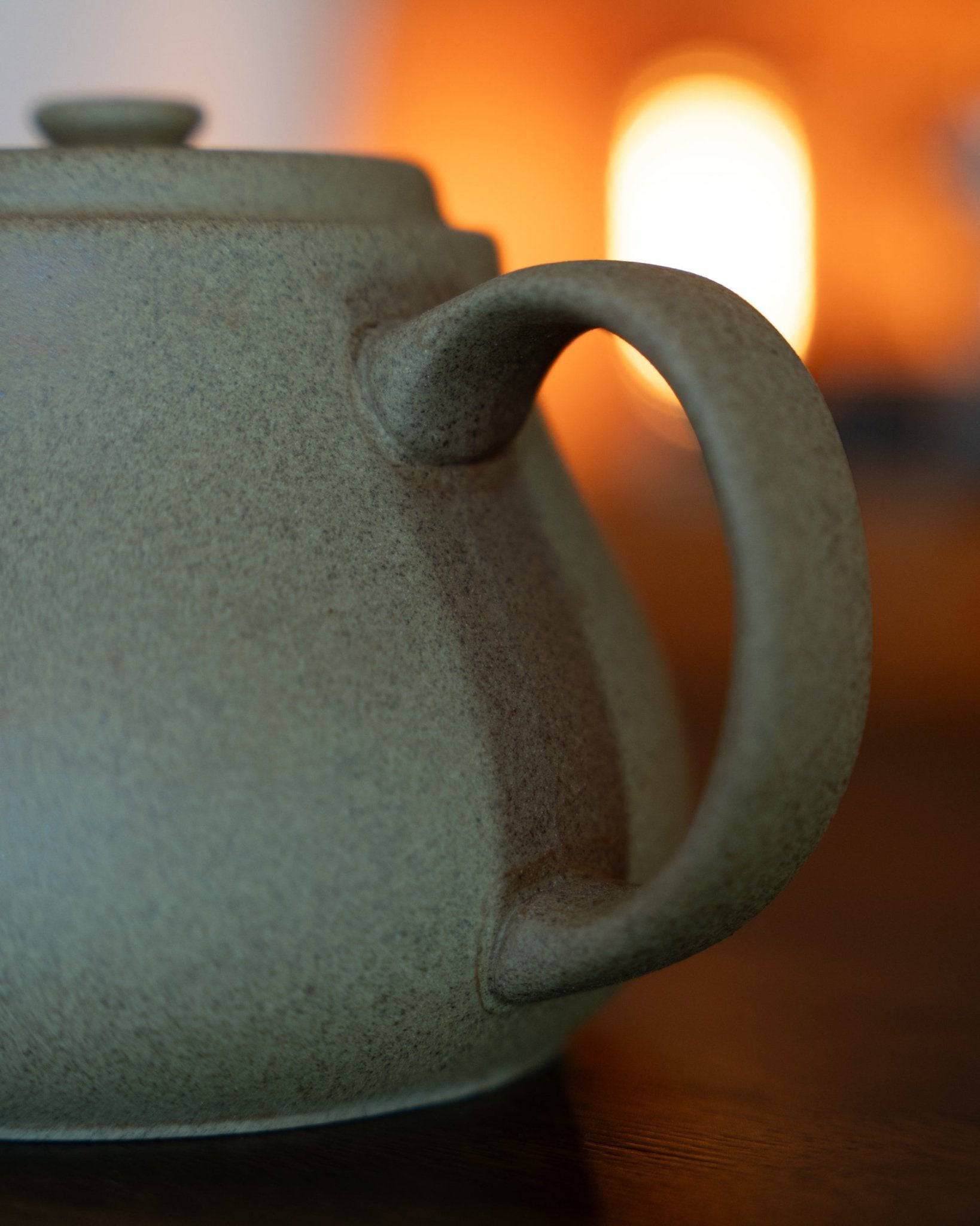 Close-up of Kalita & HASAMI sandstone coffee pot in matte green, showcasing textured surface and curved handle against warm, blurred background light
