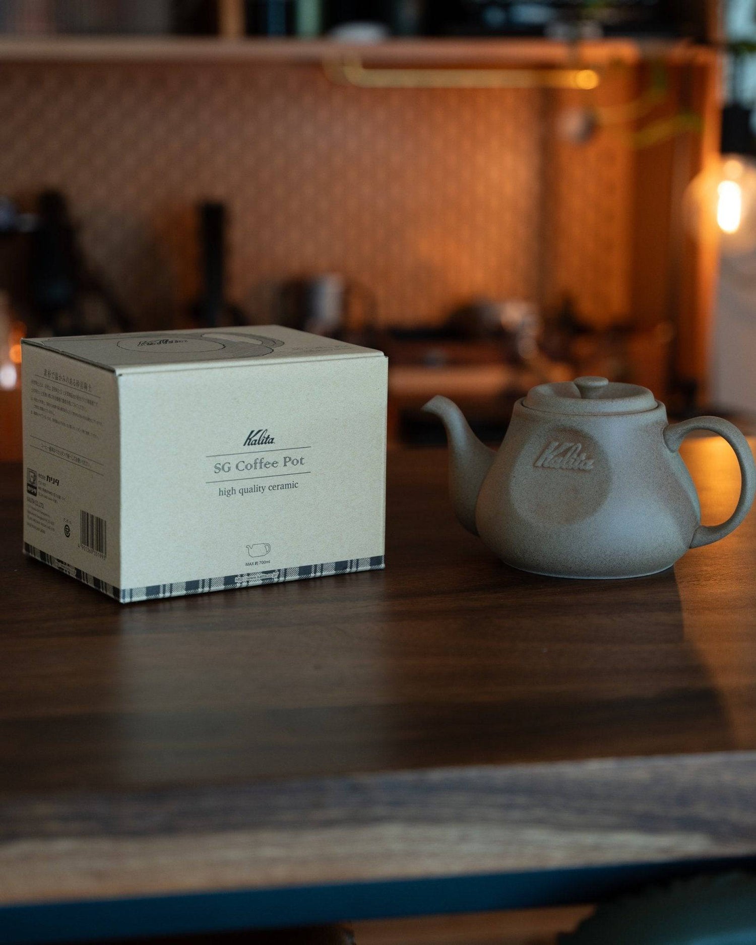 Kalita & HASAMI sandstone coffee pot with product box on wooden table, warm lighting, kitchen background. Matte gray pot showcases traditional Japanese craftsmanship blended with modern design.