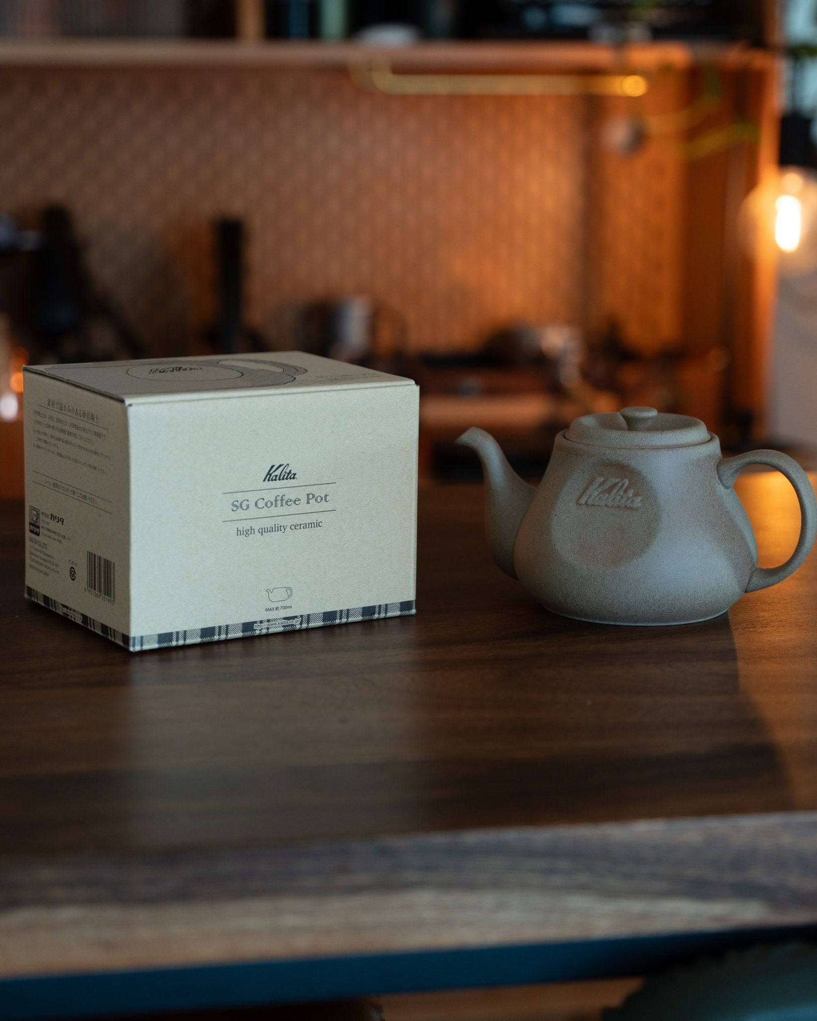 Kalita & HASAMI sandstone coffee pot with product box on wooden table, warm lighting, kitchen background. Matte gray pot showcases traditional Japanese craftsmanship blended with modern design.