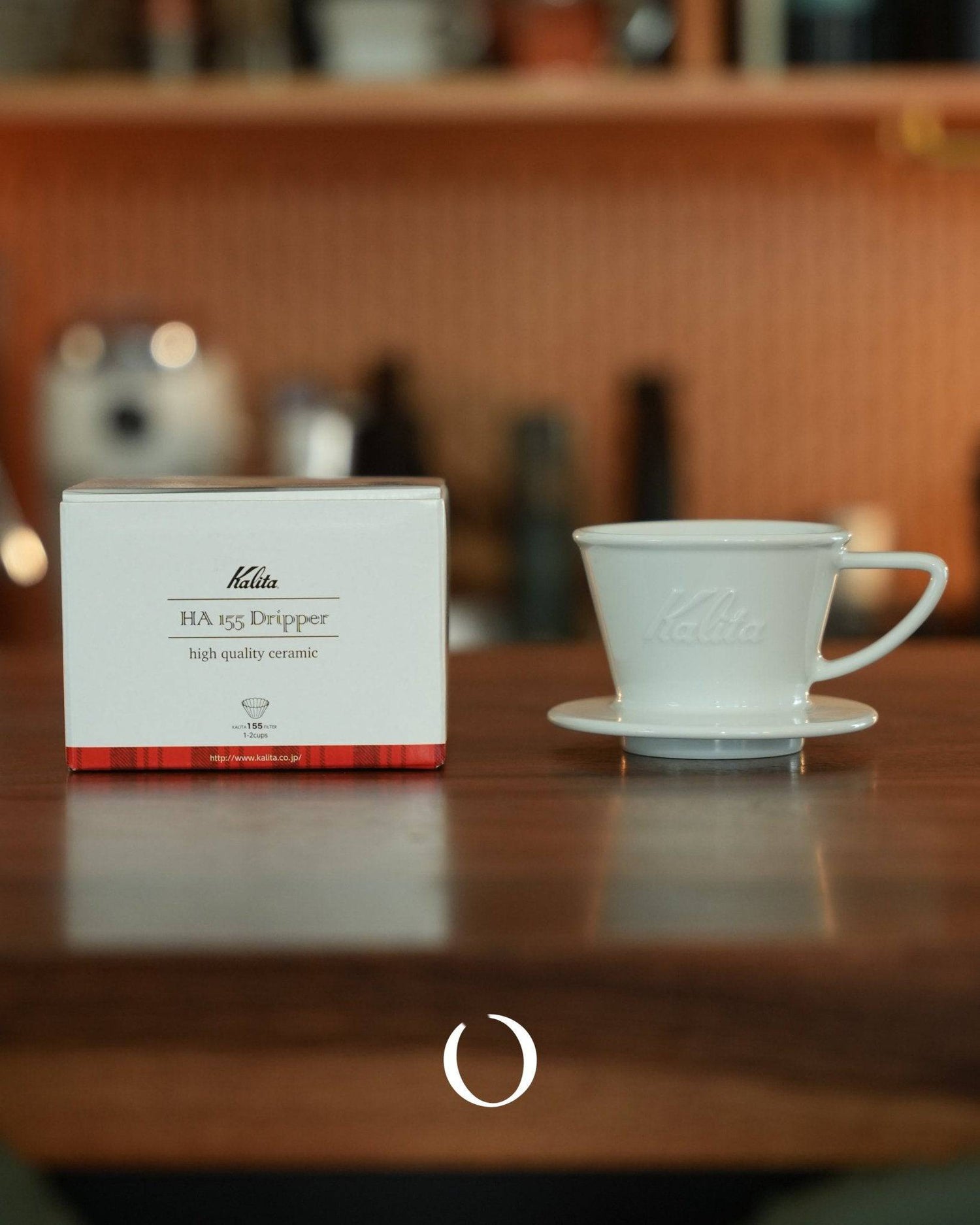 Kalita HA-155 ceramic coffee dripper with product box on wooden table, white porcelain pour-over cone and saucer, warm background with coffee equipment