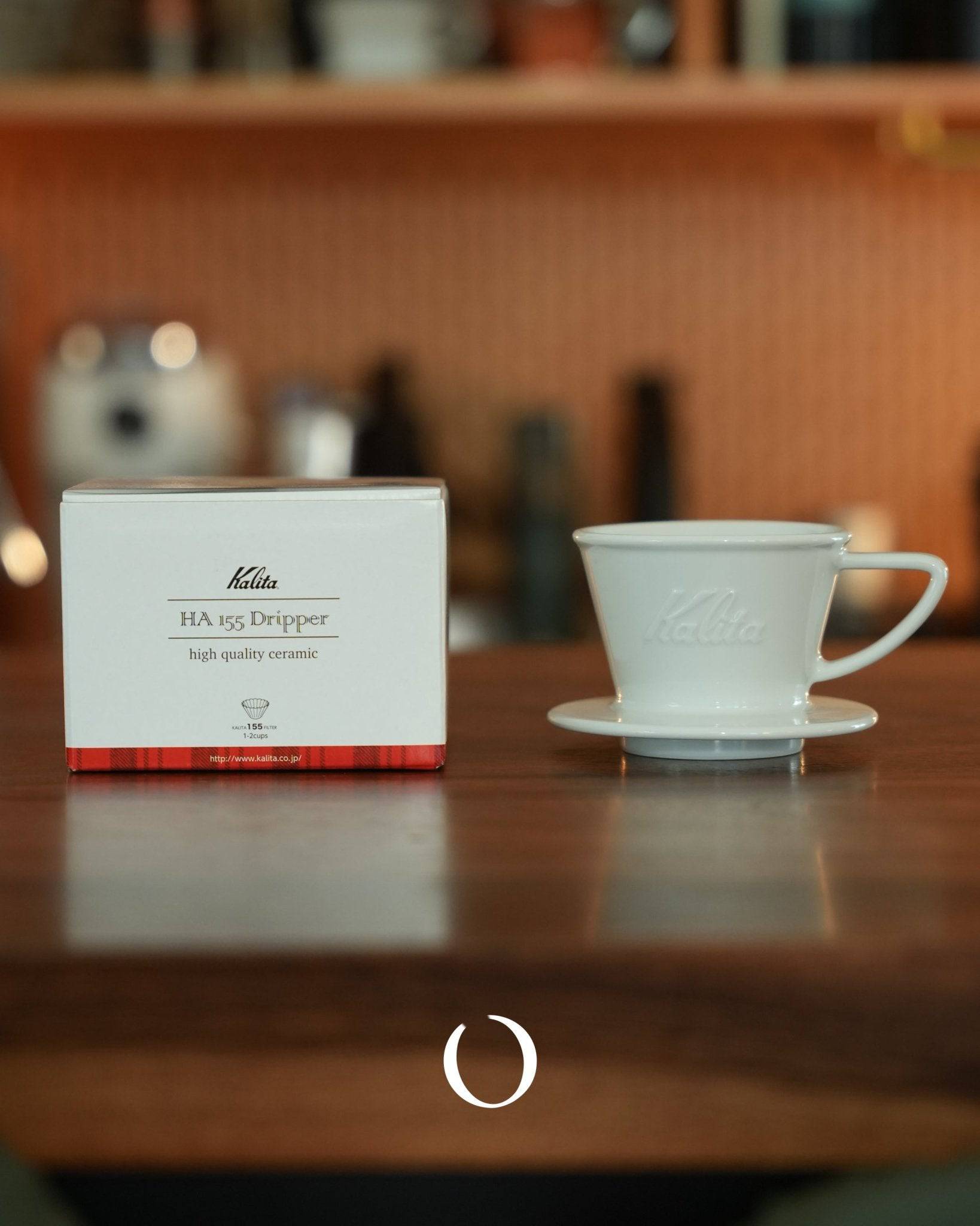 Kalita HA-155 ceramic coffee dripper with product box on wooden table, white porcelain pour-over cone and saucer, warm background with coffee equipment