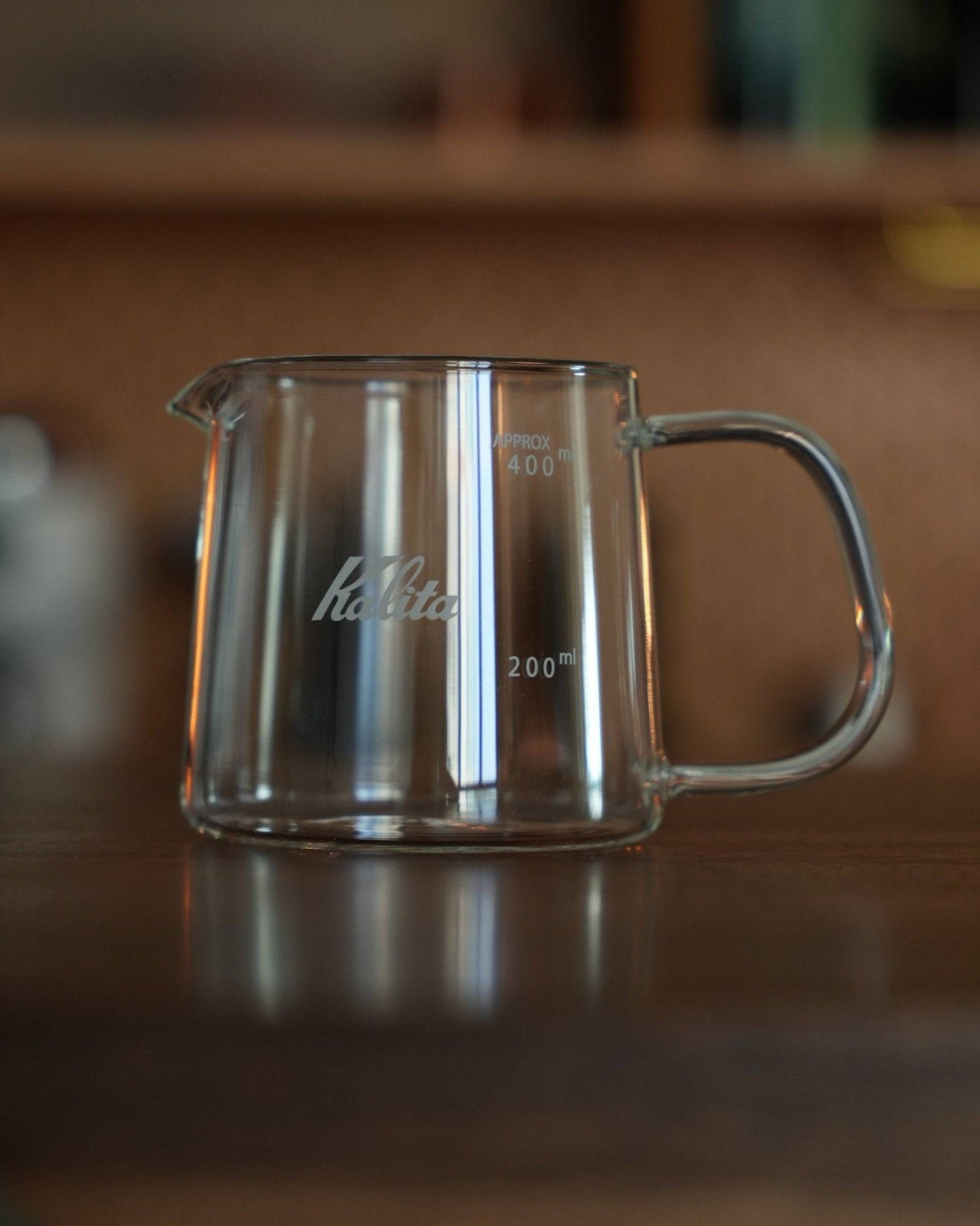 Kalita JUG400 heat-resistant glass coffee jug, 400ml capacity, with clear measurement markings and Kalita branding, featuring a sturdy handle and spout for precise pouring, displayed on a wooden surface in a warm, inviting setting