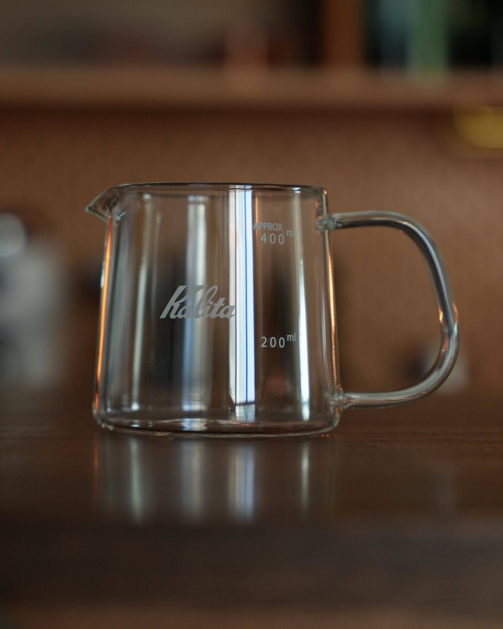 Kalita JUG400 heat-resistant glass coffee jug, 400ml capacity, with clear measurement markings and Kalita branding, featuring a sturdy handle and spout for precise pouring, displayed on a wooden surface in a warm, inviting setting