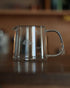 Kalita JUG400 heat-resistant glass coffee jug, 400ml capacity, with clear measurement markings and Kalita branding, featuring a sturdy handle and spout for precise pouring, displayed on a wooden surface in a warm, inviting setting