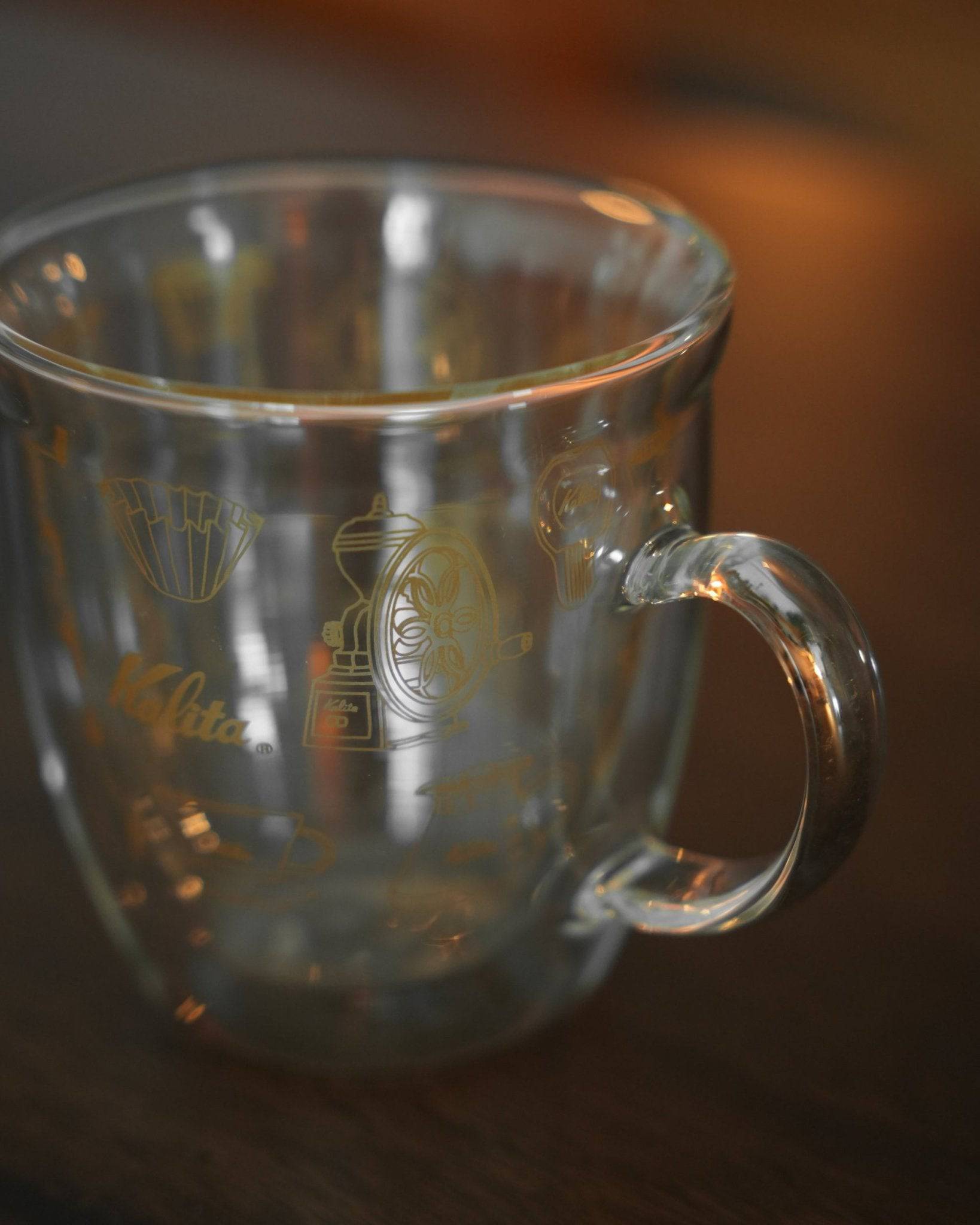 Kalita double-walled glass mug with gold coffee grinder and filter design, showcasing transparent construction and curved handle on wooden surface