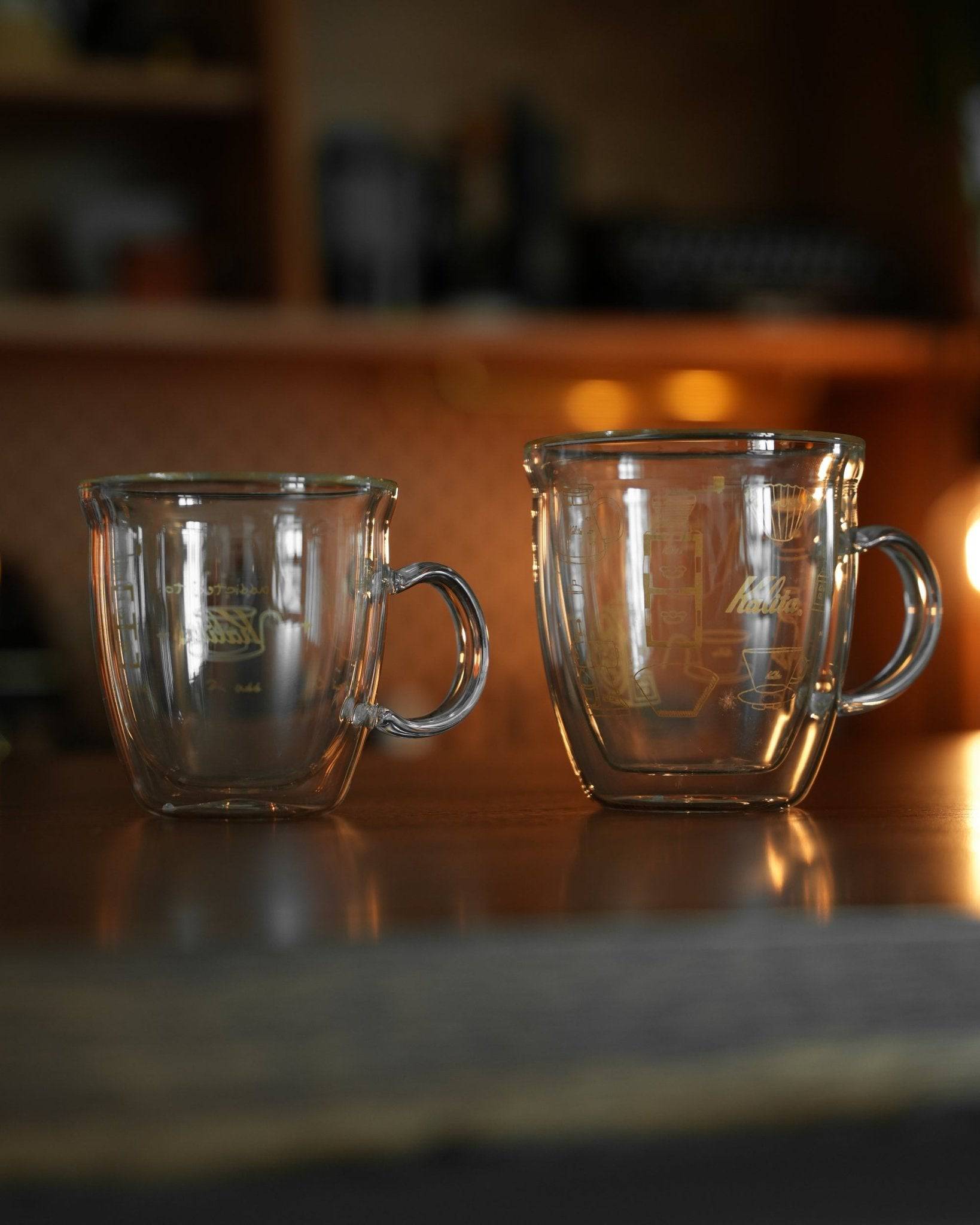 Two Kalita double-walled glass coffee mugs on wooden surface, one smaller and one larger, with Kalita logo visible on the larger mug. Warm ambient lighting in background.