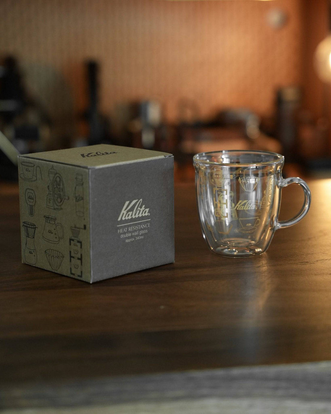 Kalita double-walled glass mug with product box on wooden surface, coffee equipment in background. Clear mug showcases Kalita logo and unique design. Packaging features coffee gear illustrations.
