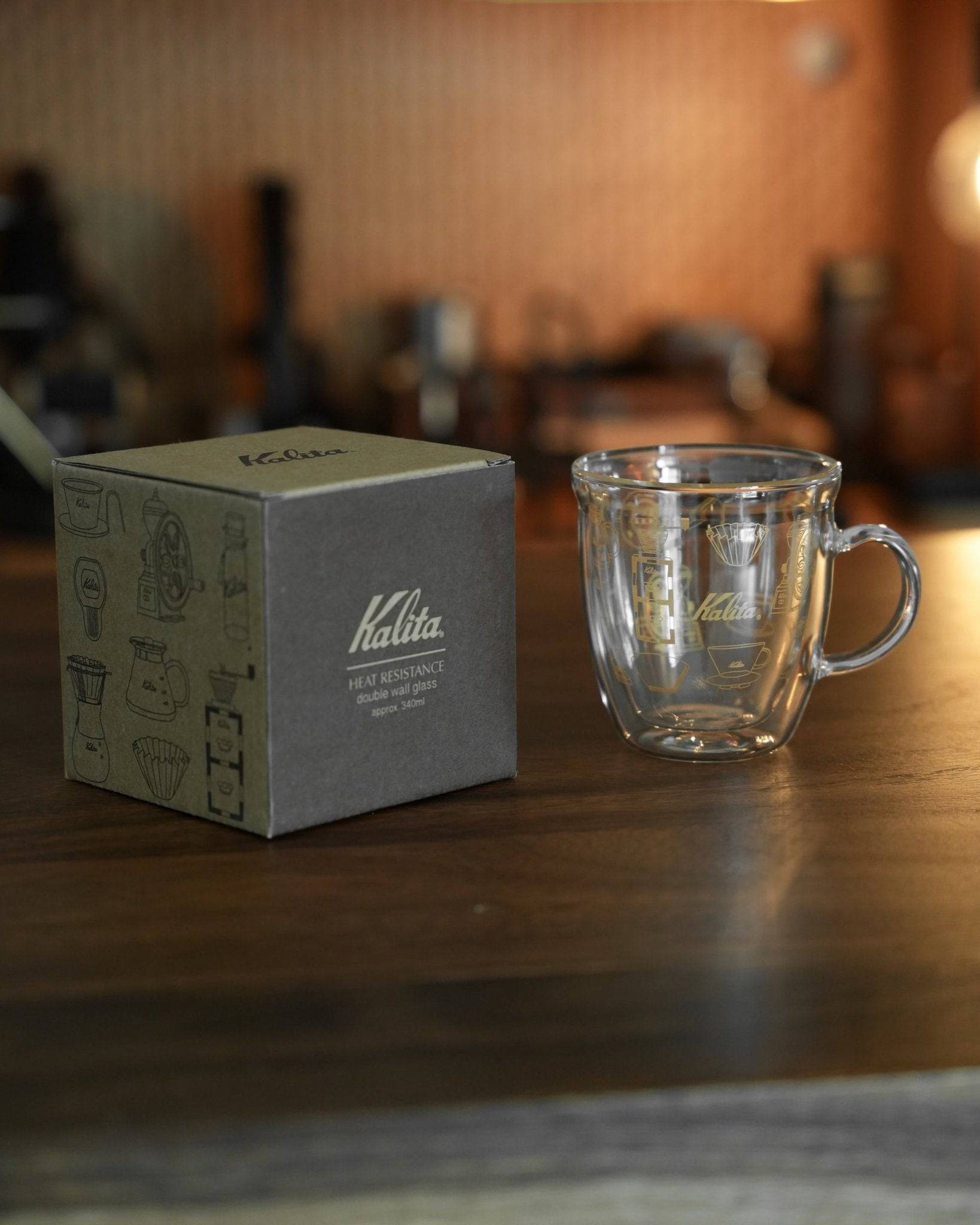 Kalita double-walled glass mug with product box on wooden surface, coffee equipment in background. Clear mug showcases Kalita logo and unique design. Packaging features coffee gear illustrations.